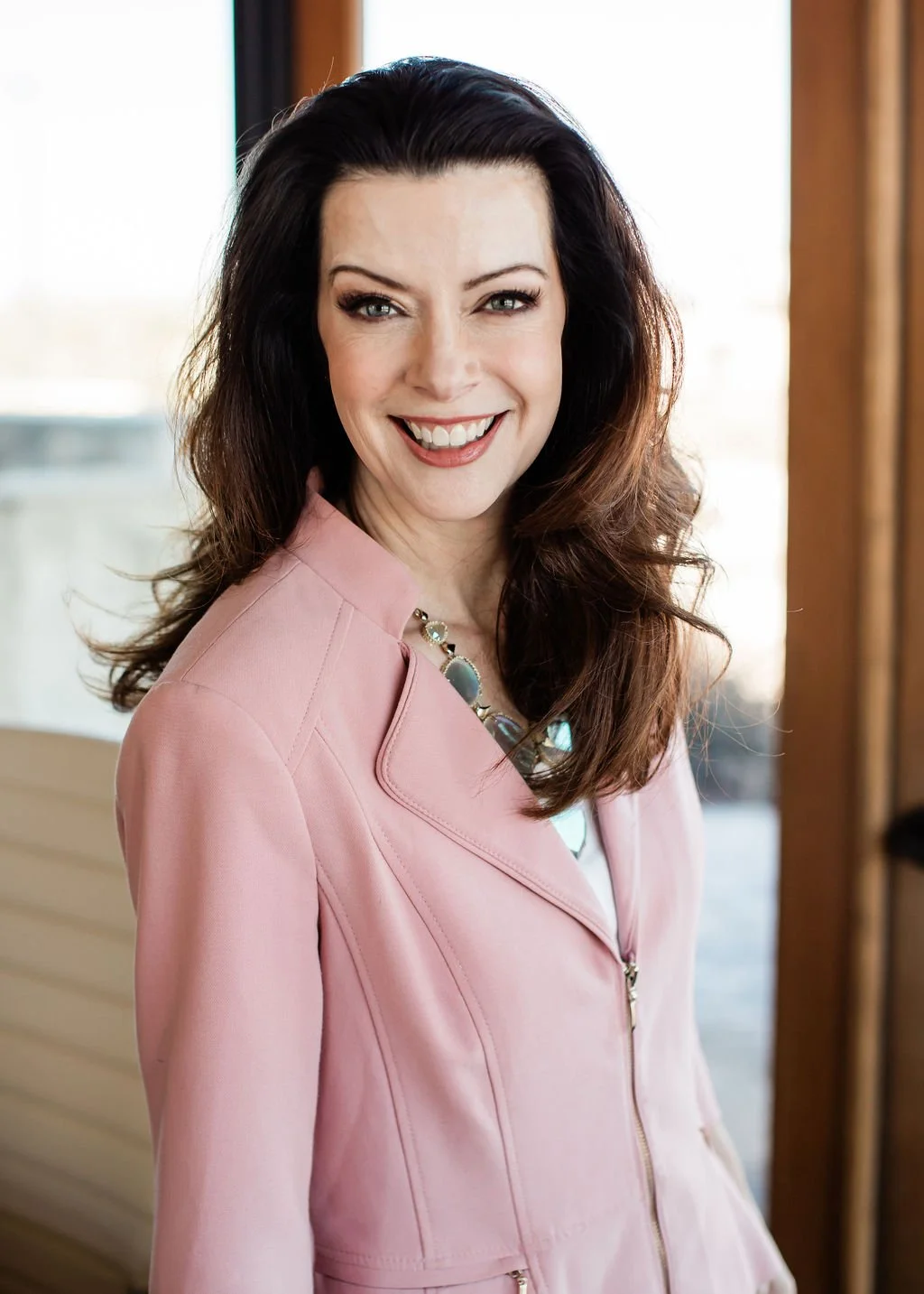 A woman with long dark hair, smiling, wearing a light pink blazer and a statement necklace, standing indoors near a window.