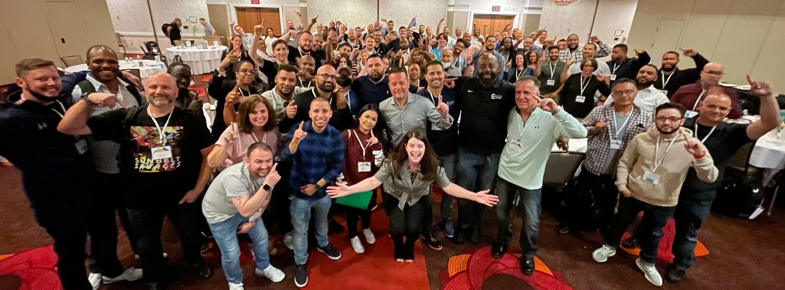 Large group of diverse people gathered in a conference room, smiling, some holding up number one fingers, celebrating together.
