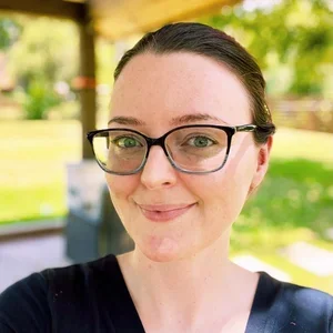 A woman with dark hair, glasses, and a black shirt, smiling outdoors in a park with trees and greenery in the background.