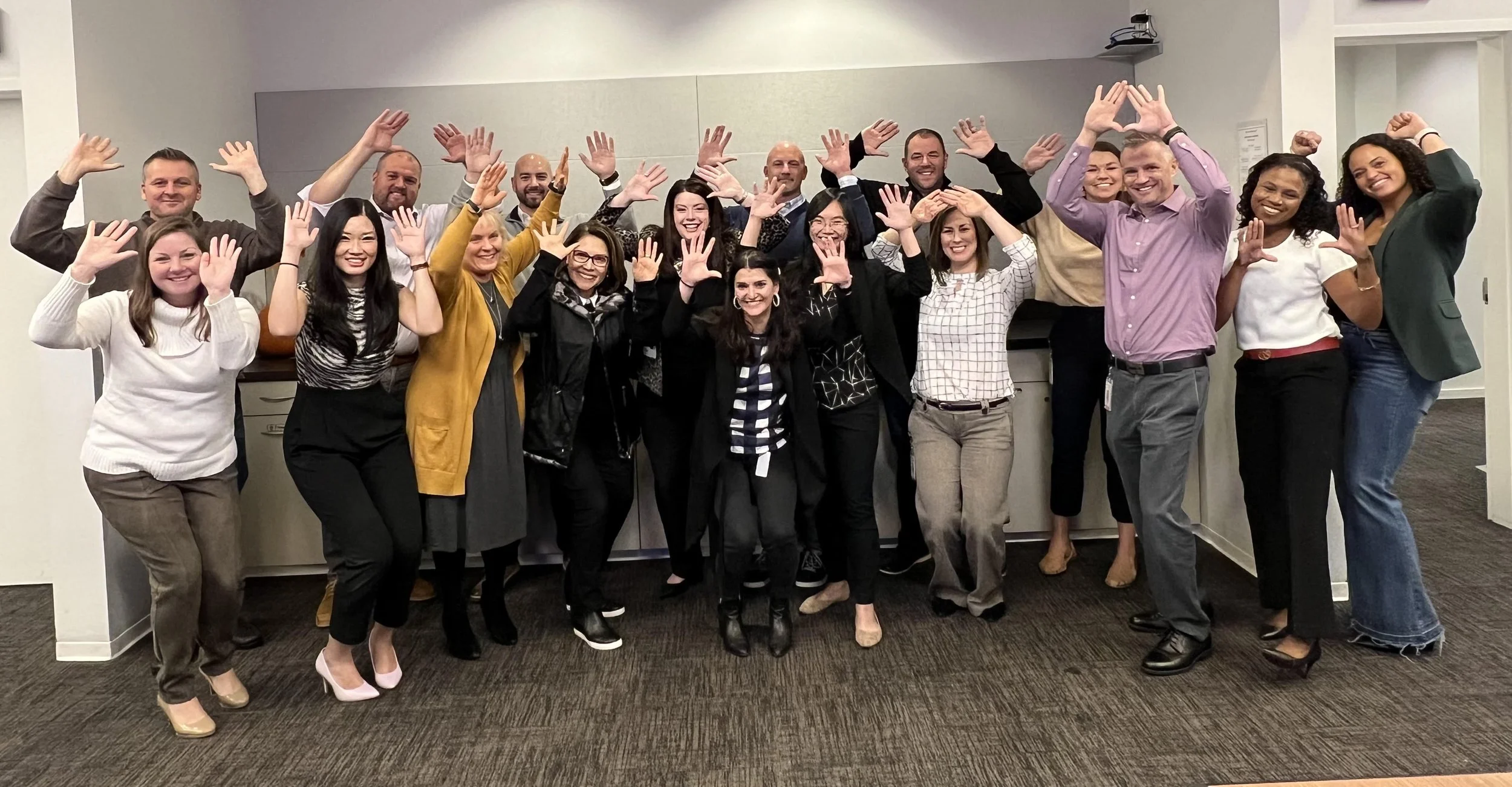 A group of 17 diverse smiling people posing together in an office, raising their hands in celebration or greeting.