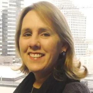 Close-up of a woman with shoulder-length blond hair, smiling, in front of a cityscape with tall buildings.