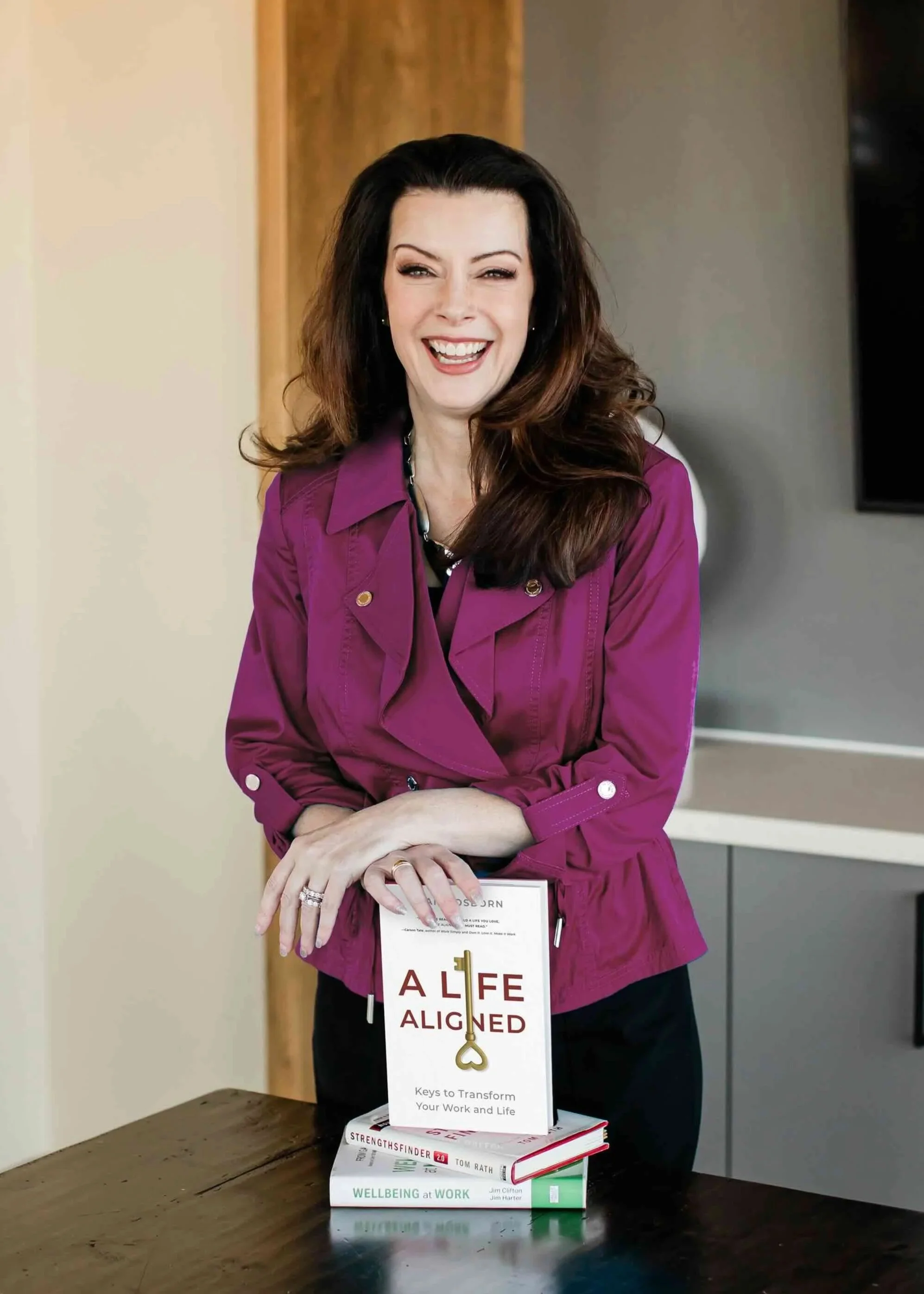 A woman with long brown hair smiling, wearing a purple jacket, standing behind a table with books stacked on it, one titled 'ALIFE ALIGNED' with a key symbol on the cover.