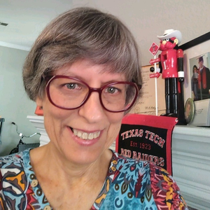 A smiling woman with short gray hair and glasses, wearing a colorful blouse, standing in a room with a fireplace decorated with a nutcracker and other festive decorations.