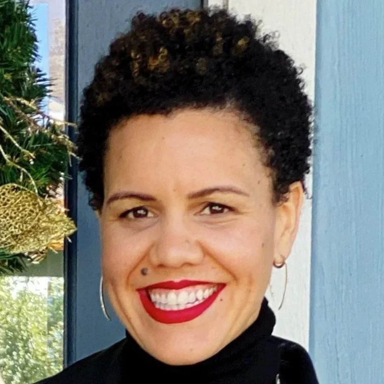 Close-up of a woman with short, curly hair, red lipstick, hoop earrings, smiling, standing outdoors near a decorated Christmas tree.