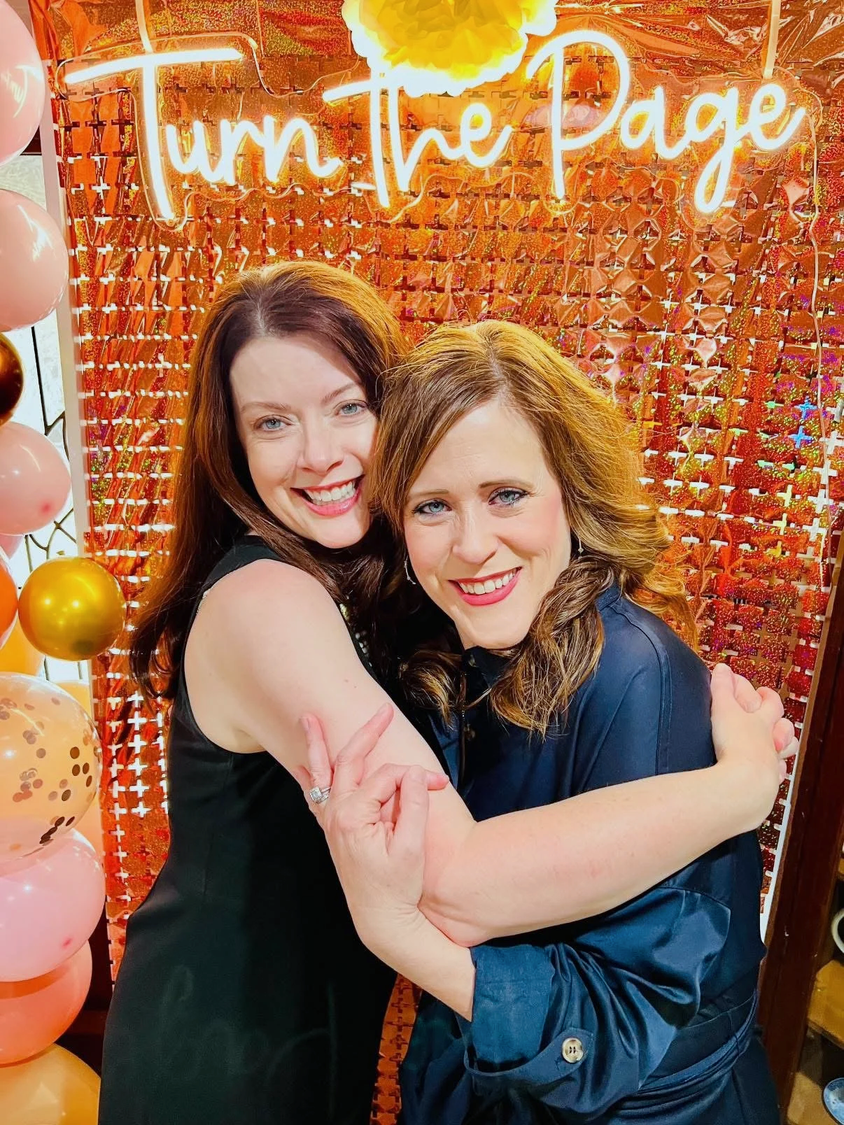 Two women hugging each other in front of a red and pink balloon backdrop with a neon sign that says 'Turn the Page.'