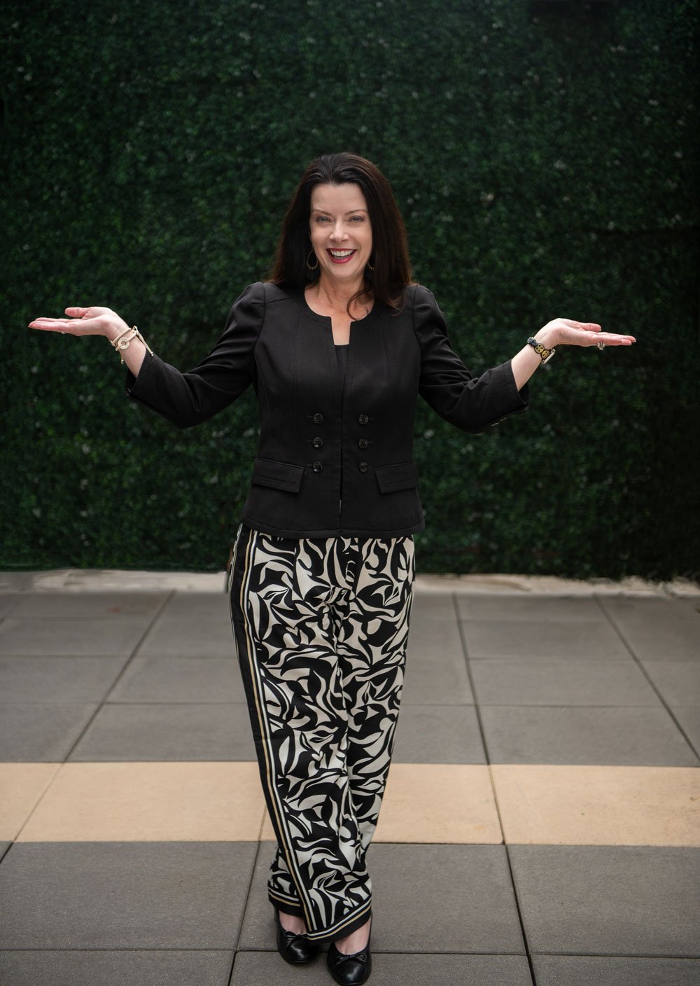 A woman with dark hair wearing a black blazer and patterned black and white pants, standing outdoors in front of a green hedge, smiling and holding her arms out to the sides.
