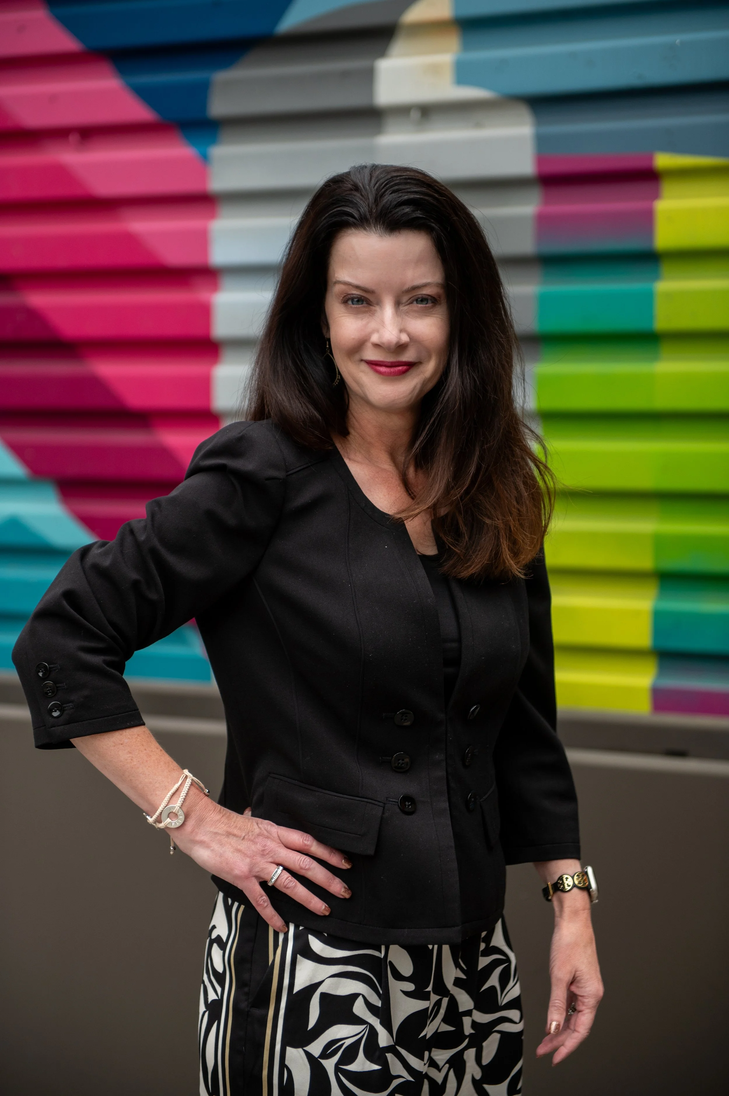 Woman with dark hair in a black blazer and patterned pants standing in front of a colorful mural.