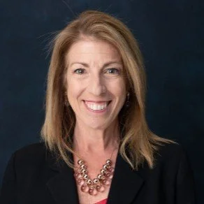 A woman with shoulder-length blonde hair smiling, wearing a black blazer and a statement necklace against a dark background.