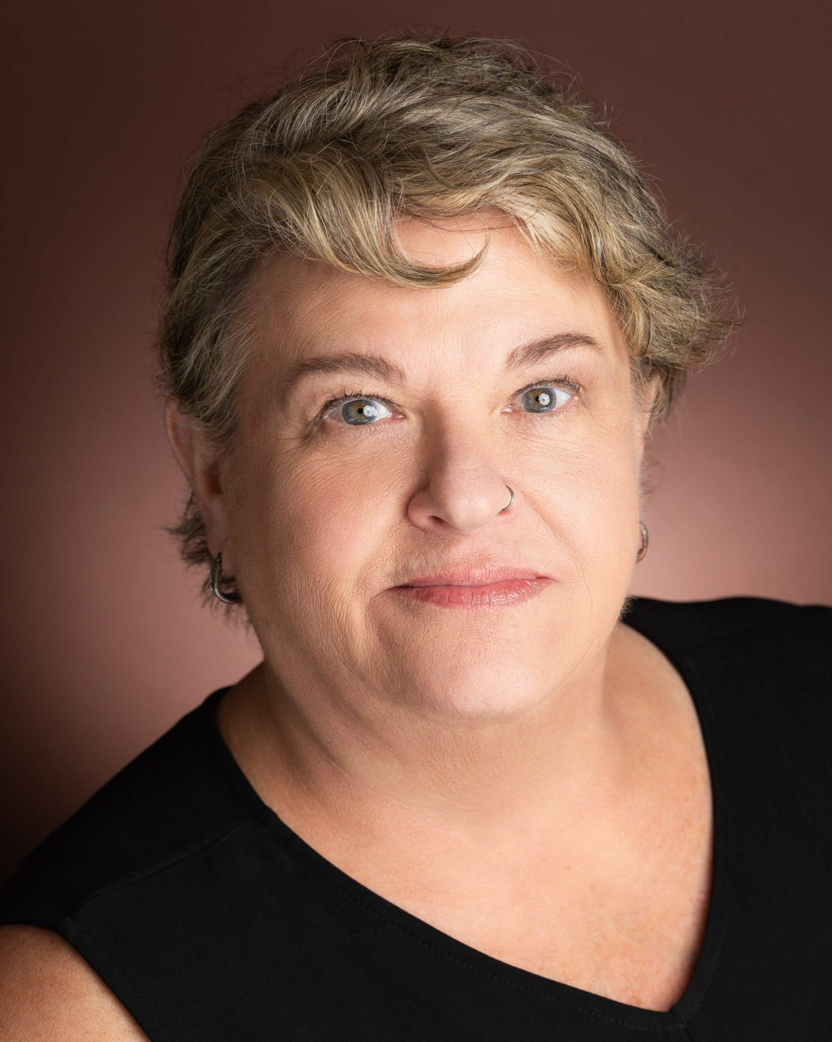 Portrait of a middle-aged woman with short, light-colored hair, blue eyes, and a nose ring, wearing a black top, against a brown background.