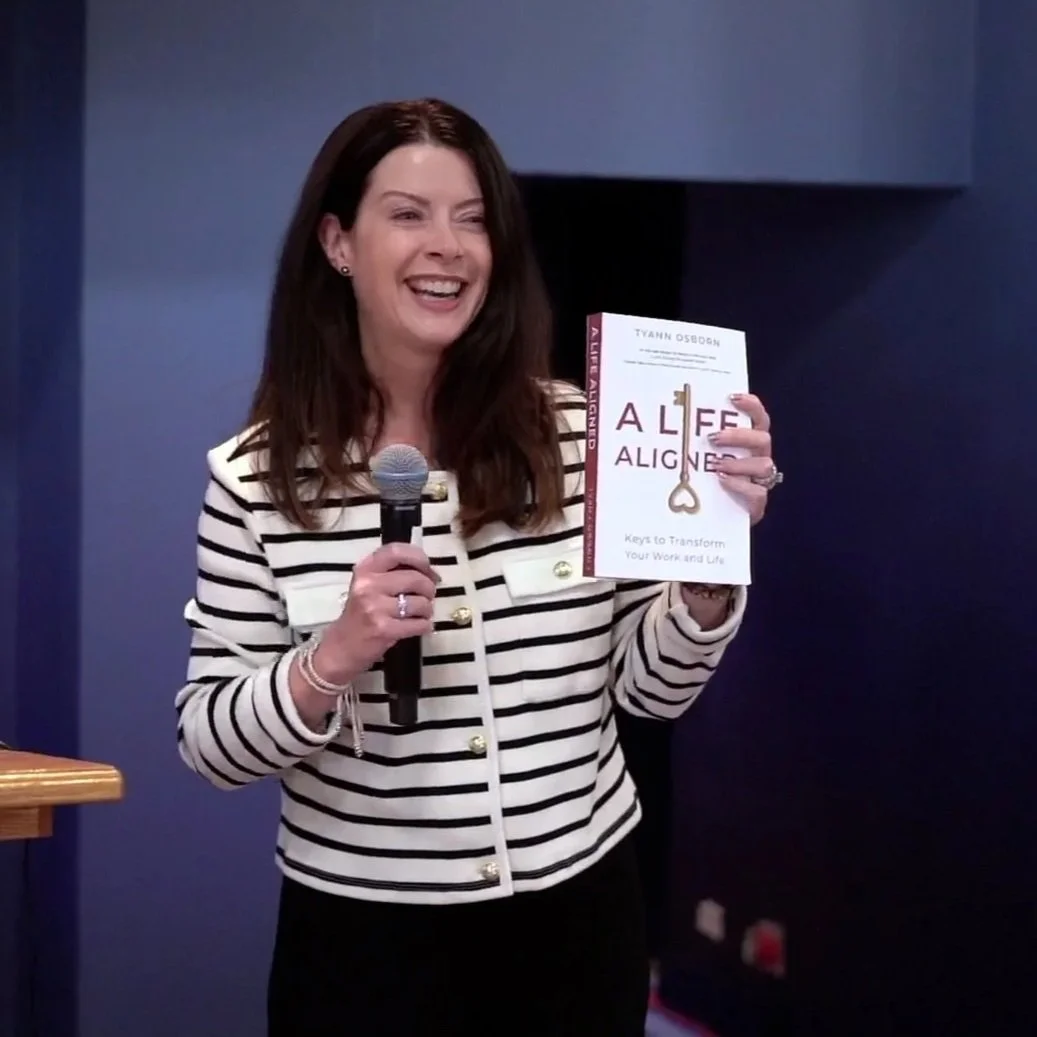 A woman with long dark hair wearing a black and white striped blazer holding a book titled 'ALIGNED' in one hand and a microphone in the other, smiling during a presentation.