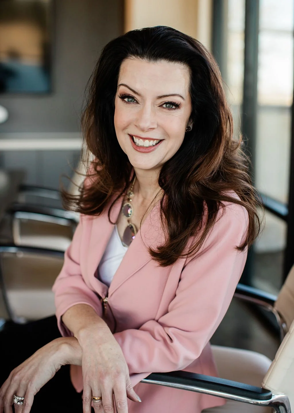 A smiling woman with long dark hair and blue eyes, wearing a pink blazer and a statement necklace, sitting in a modern office setting.