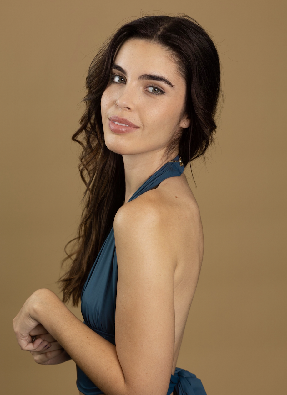 A young woman with long dark hair, wearing a blue halter dress, looking at the camera with a slight smile against a plain light brown background.