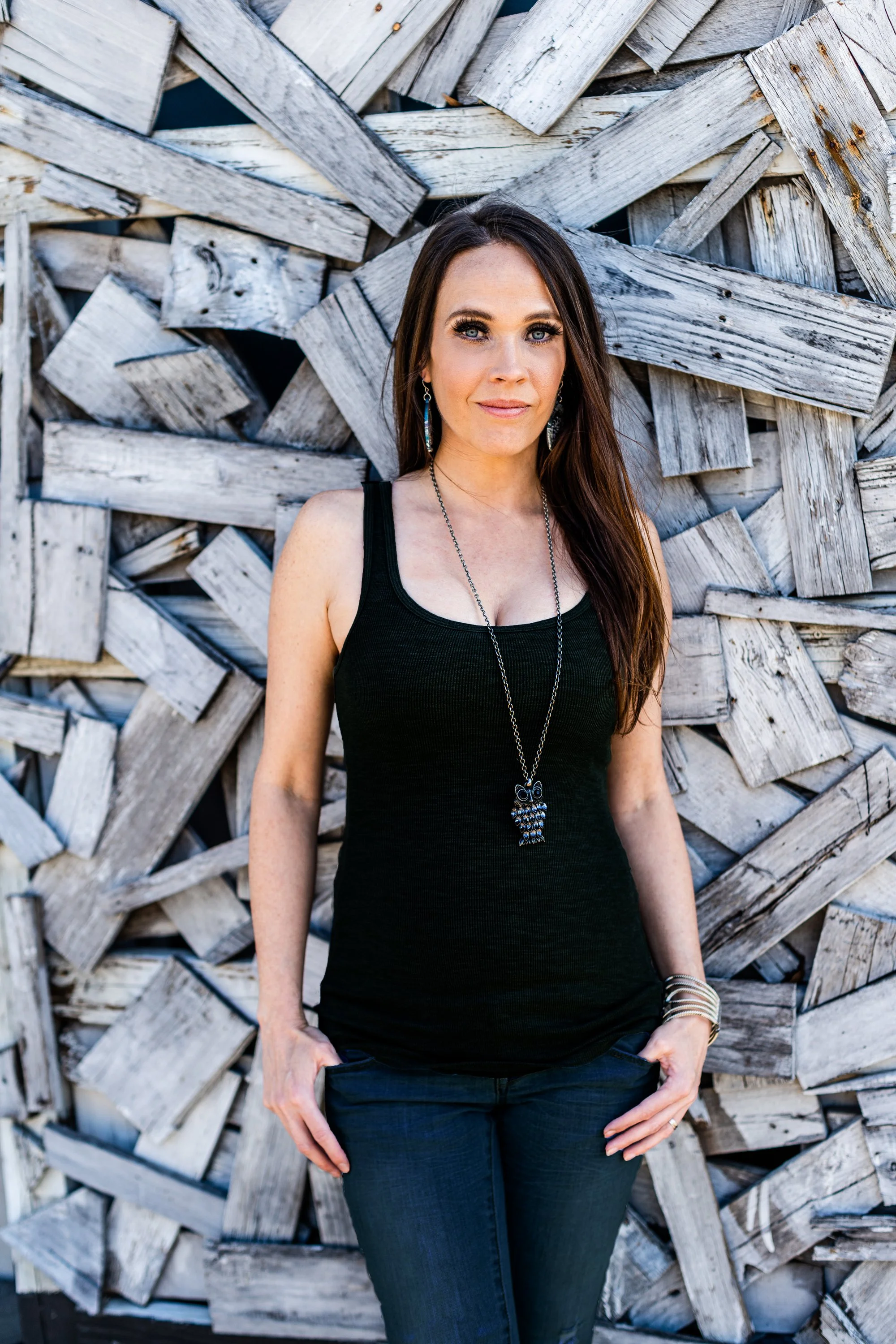 A woman with long dark hair and blue eyes wearing a black tank top, blue jeans, and a owl-shaped necklace standing in front of a weathered wood fence.