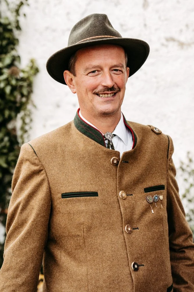 Ein Mann trägt traditionelle bayerische Kleidung, einschließlich Lederjacke, Hut und Schmuck, und lächelt in die Kamera.
