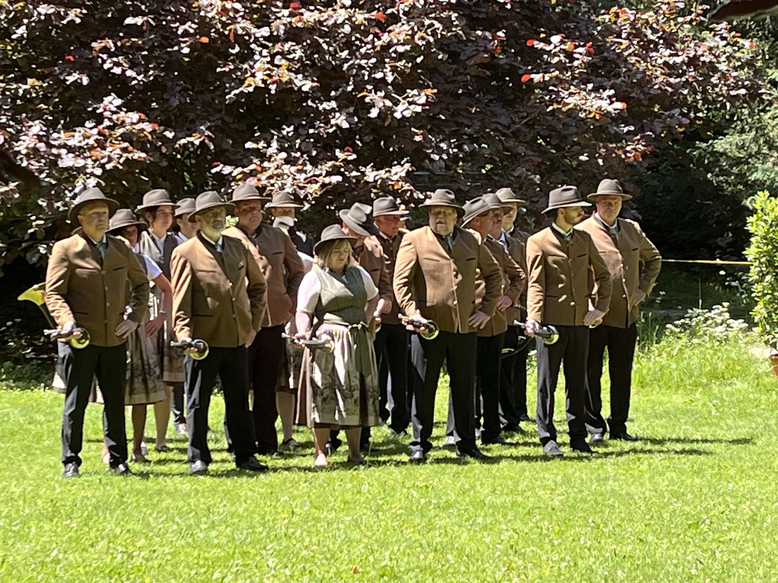 Mehrere Menschen in traditionellen Trachten und Filzhüten stehen im Garten, bei einem Jagdhornbläser-Wettbewerb.