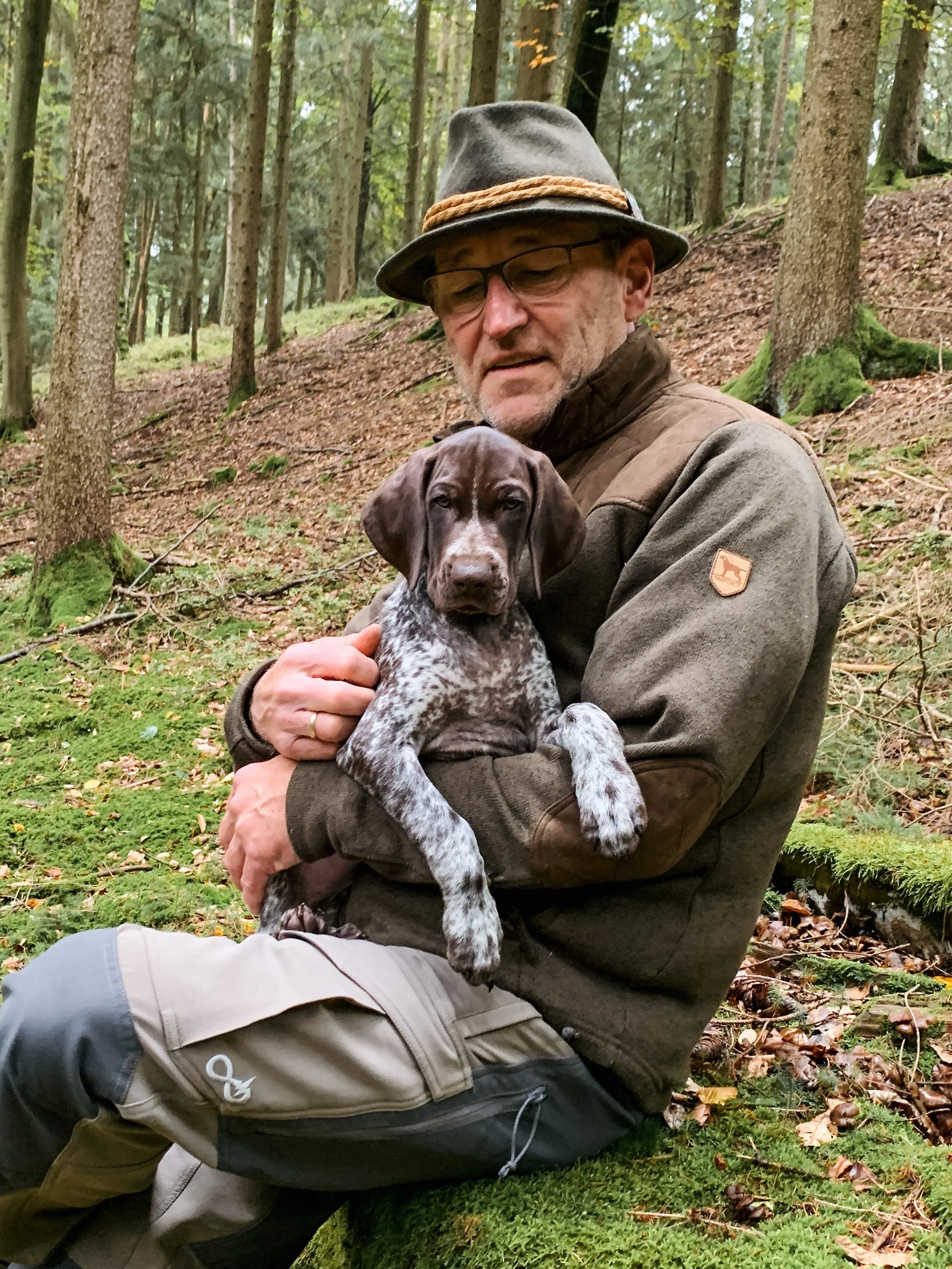 Ein älterer Mann trägt einen Filzhut, eine Brille und eine traditionelle Jagdjacke. Er steht vor einem hellen Hintergrund mit Pflanzen im Hintergrund.