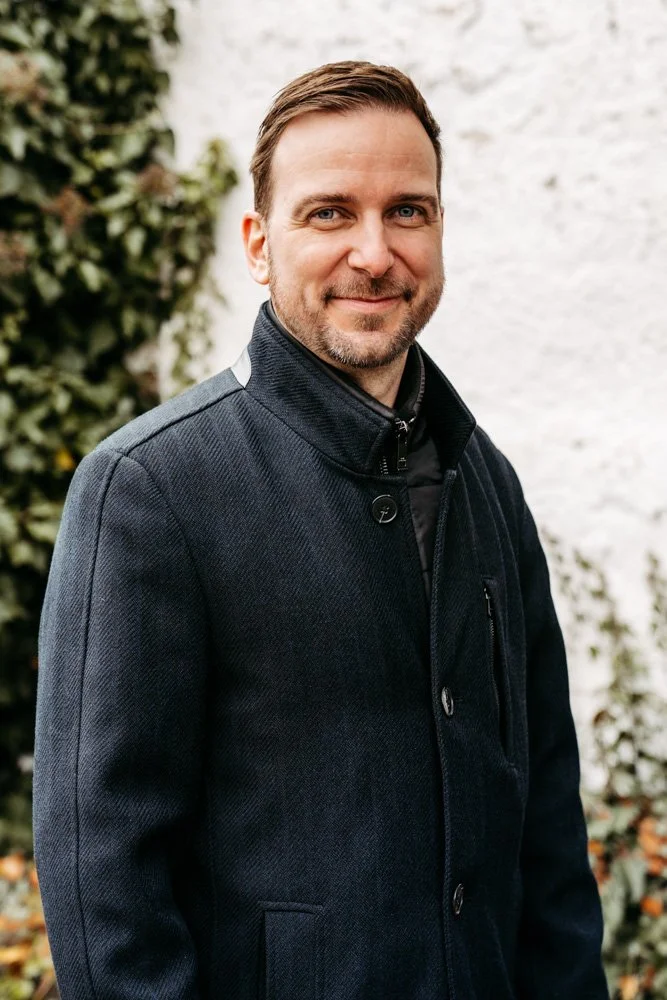 Ein Mann mit braunem Haar und Bart, trägt eine schwarze Jacke und steht vor einer weißen Mauer und grünen Pflanzen.