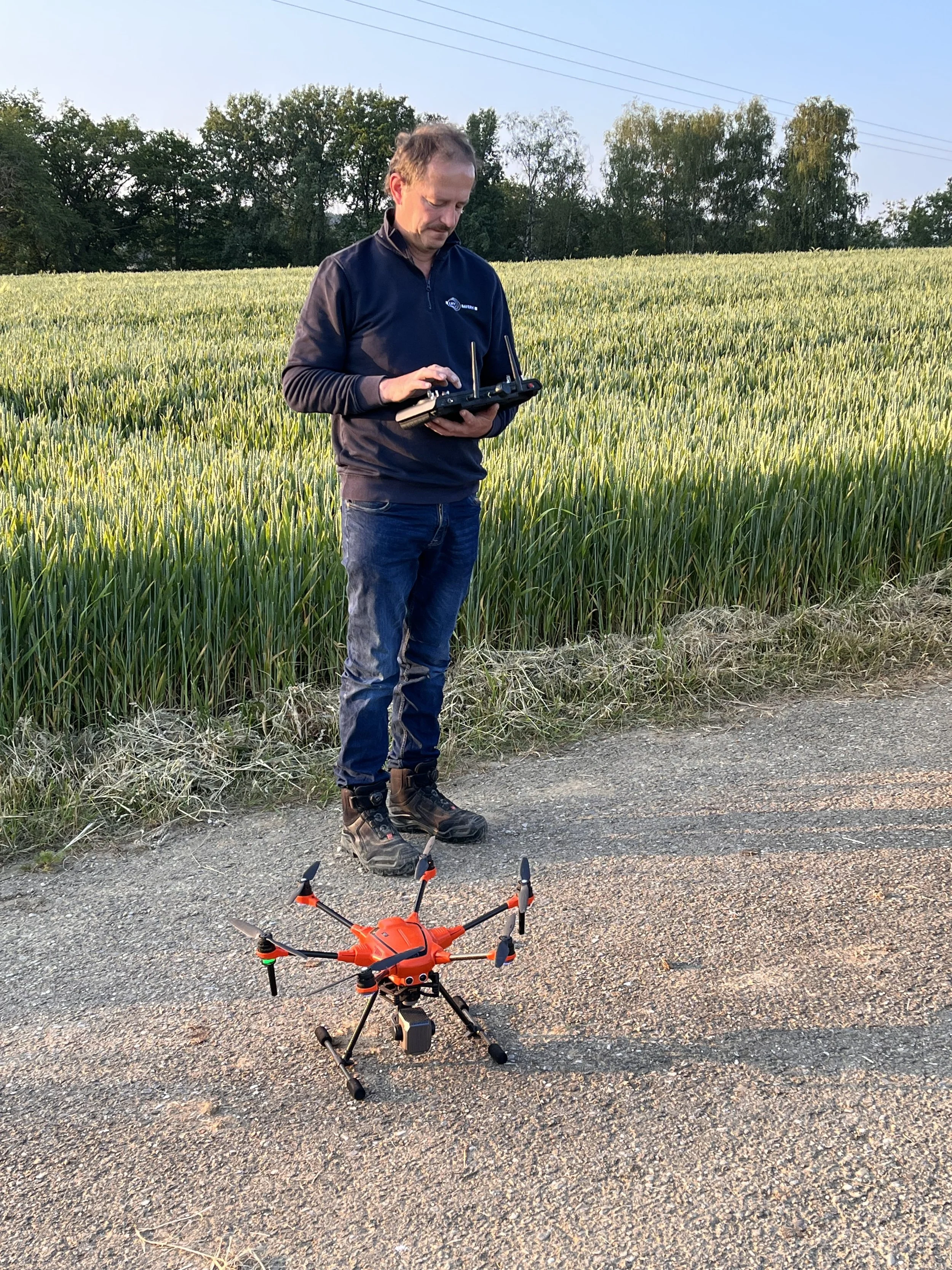 Mann steuert eine ferngesteuerte Drohne auf einer Landstraße neben einem Weizenfeld.