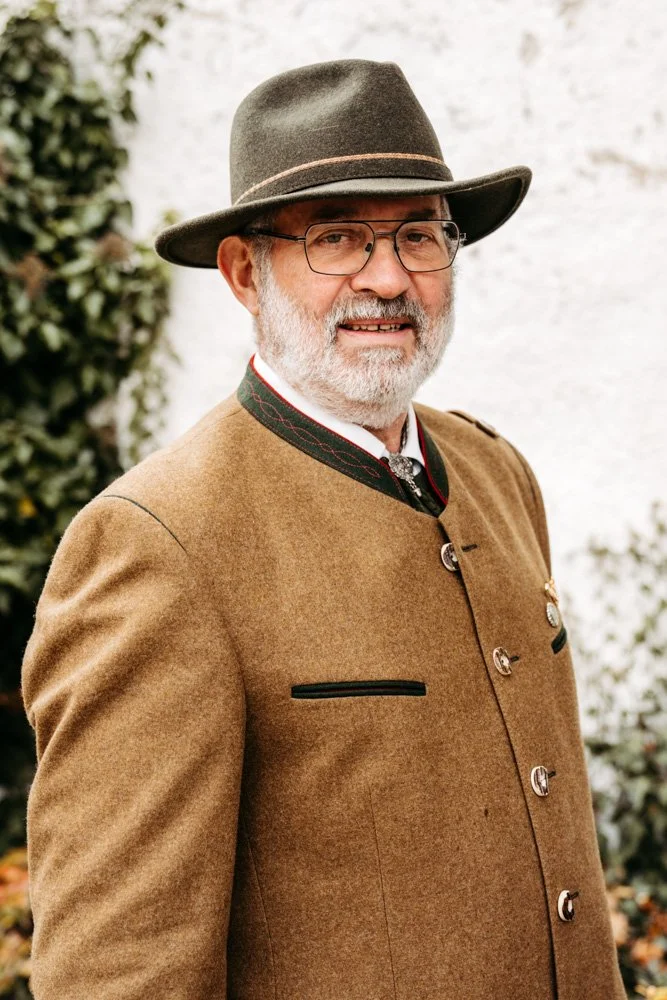 Ein Mann in traditioneller bayerischer Kleidung, trägt einen Filzhut, eine braune Jacke und Brille, steht vor einem weißen Hintergrund mit etwas Grün