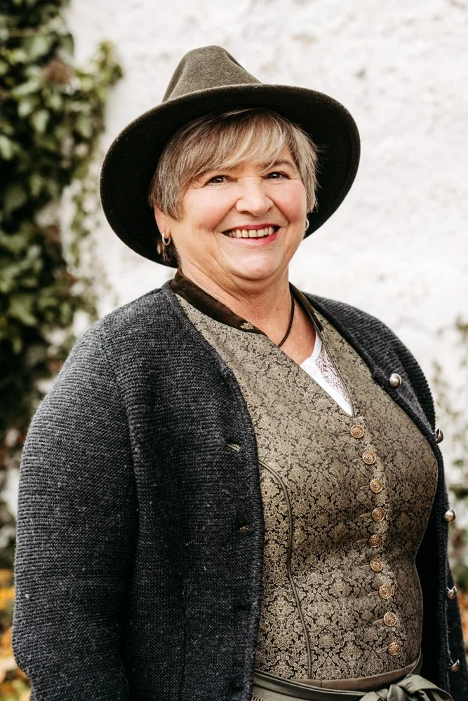 Eine lächelnde ältere Frau mit kurzen grauen Haaren trägt einen schwarzen Hut, eine graue Jacke und eine goldene, gemusterte Trachten-Bluse.