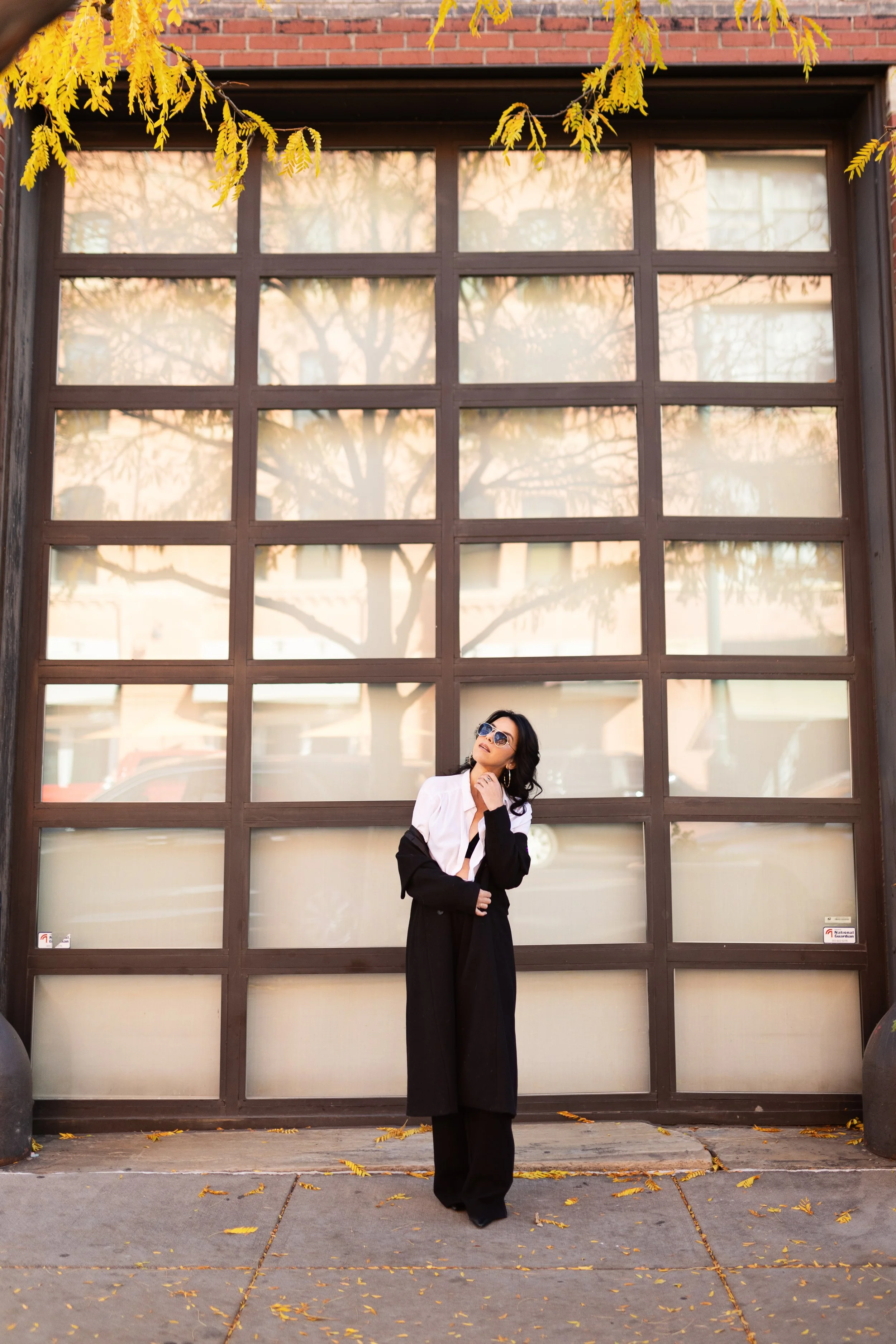 A woman wearing sunglasses and a black and white outfit stands in front of a large window with a grid pattern. She has her hand on her chin and is looking up, with yellow leaves scattered on the ground around her.