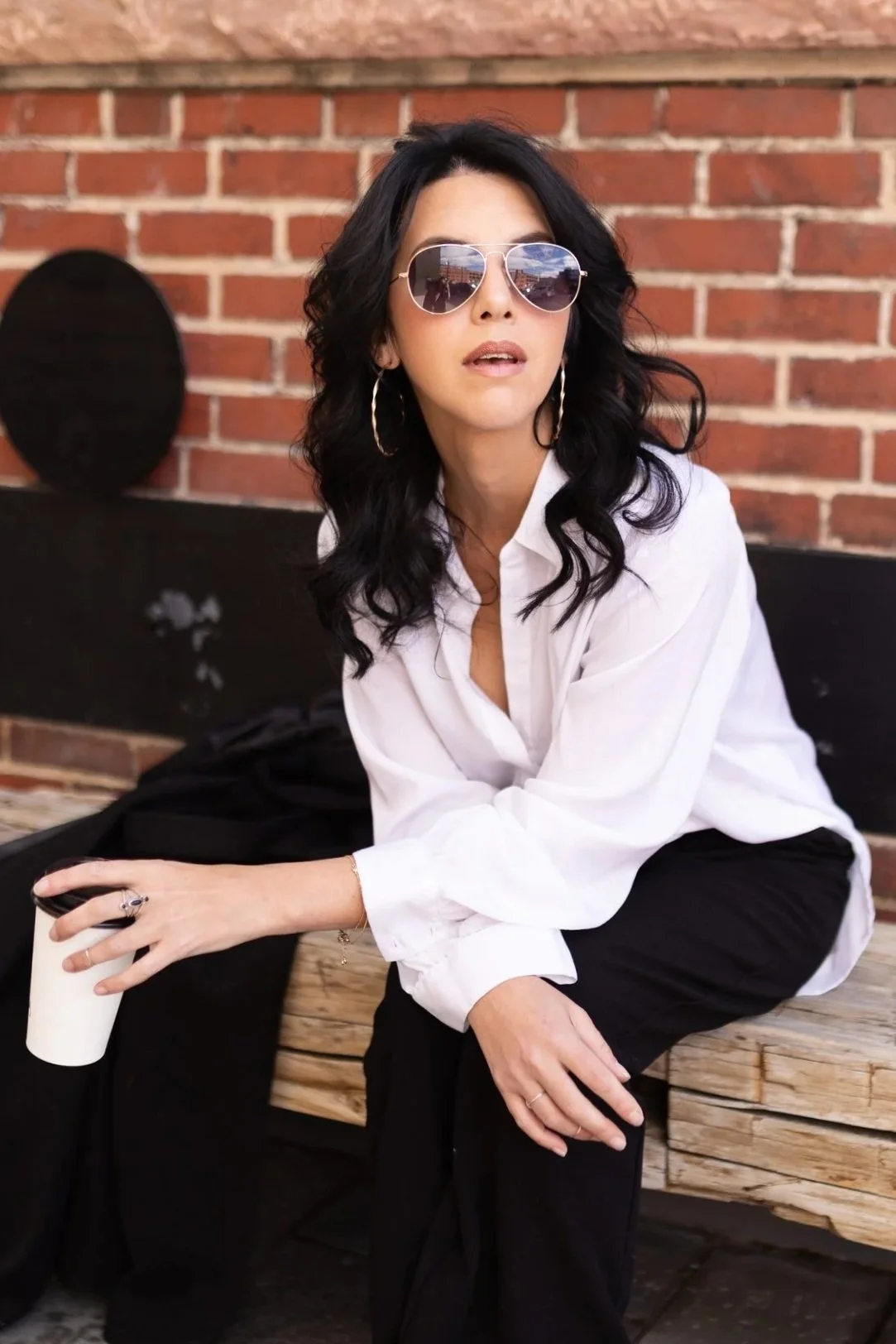 A woman with black curly hair wearing sunglasses, a white button-up shirt, and black pants sitting on a wooden bench against a brick wall, holding a white coffee cup.