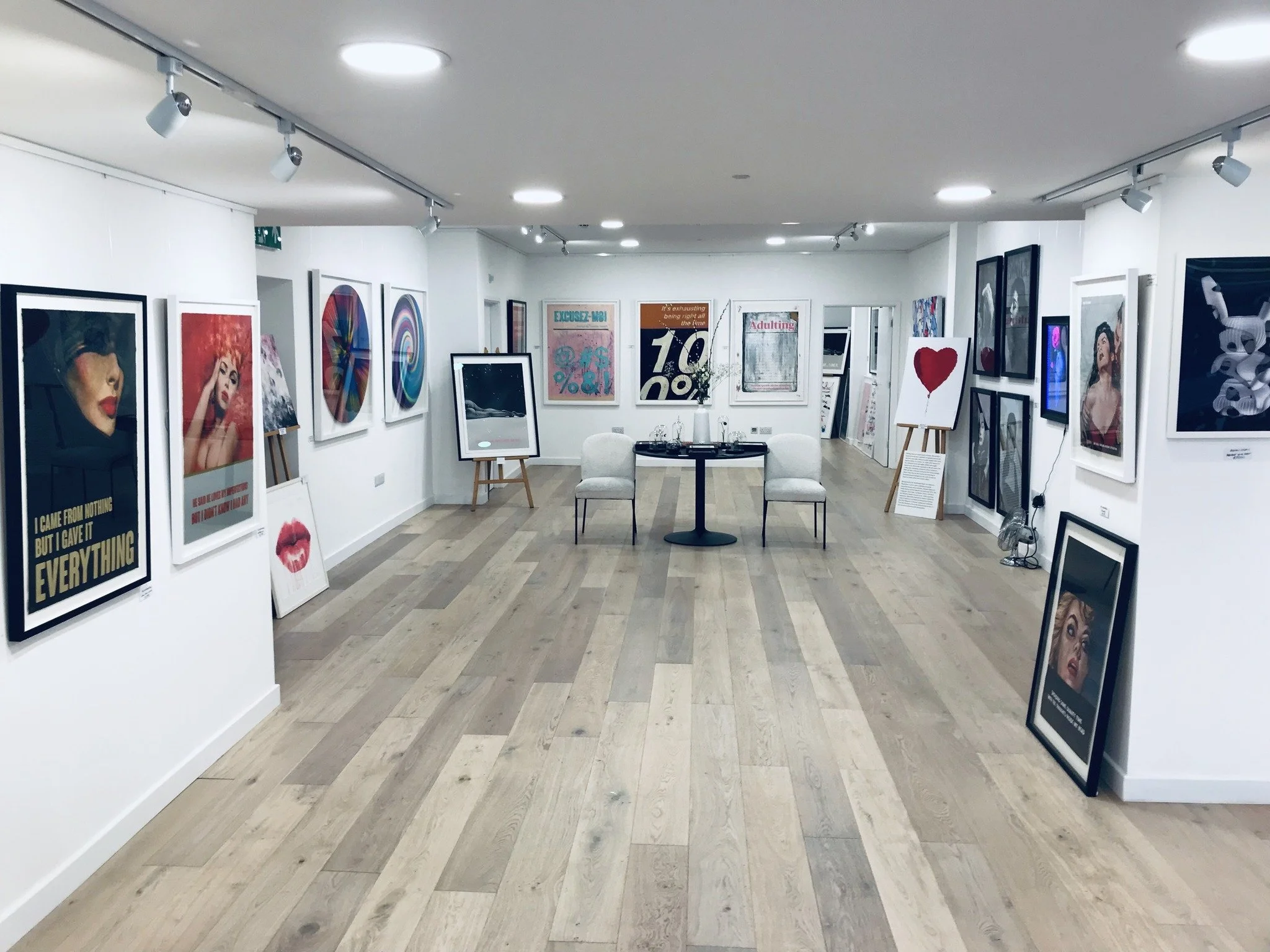 An art gallery with various framed artworks displayed on white walls, including portraits, abstract paintings, and graphic prints. There are two white chairs and a small round table in the center.