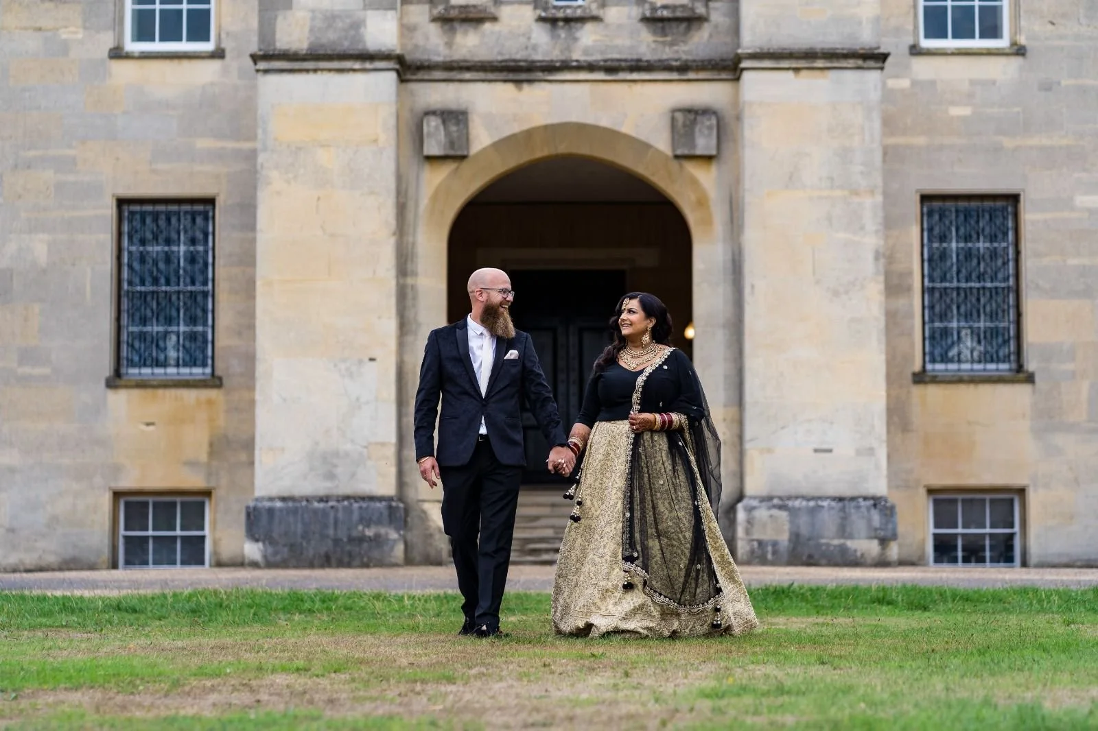 A couple holding hands and walking on grass in front of an old stone building, smiling at each other.
