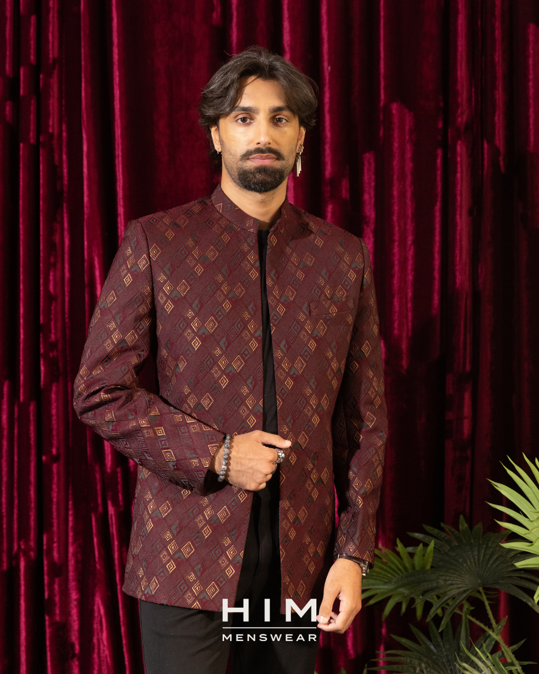 Man wearing a burgundy sherwani jacket in front of a red velvet curtain