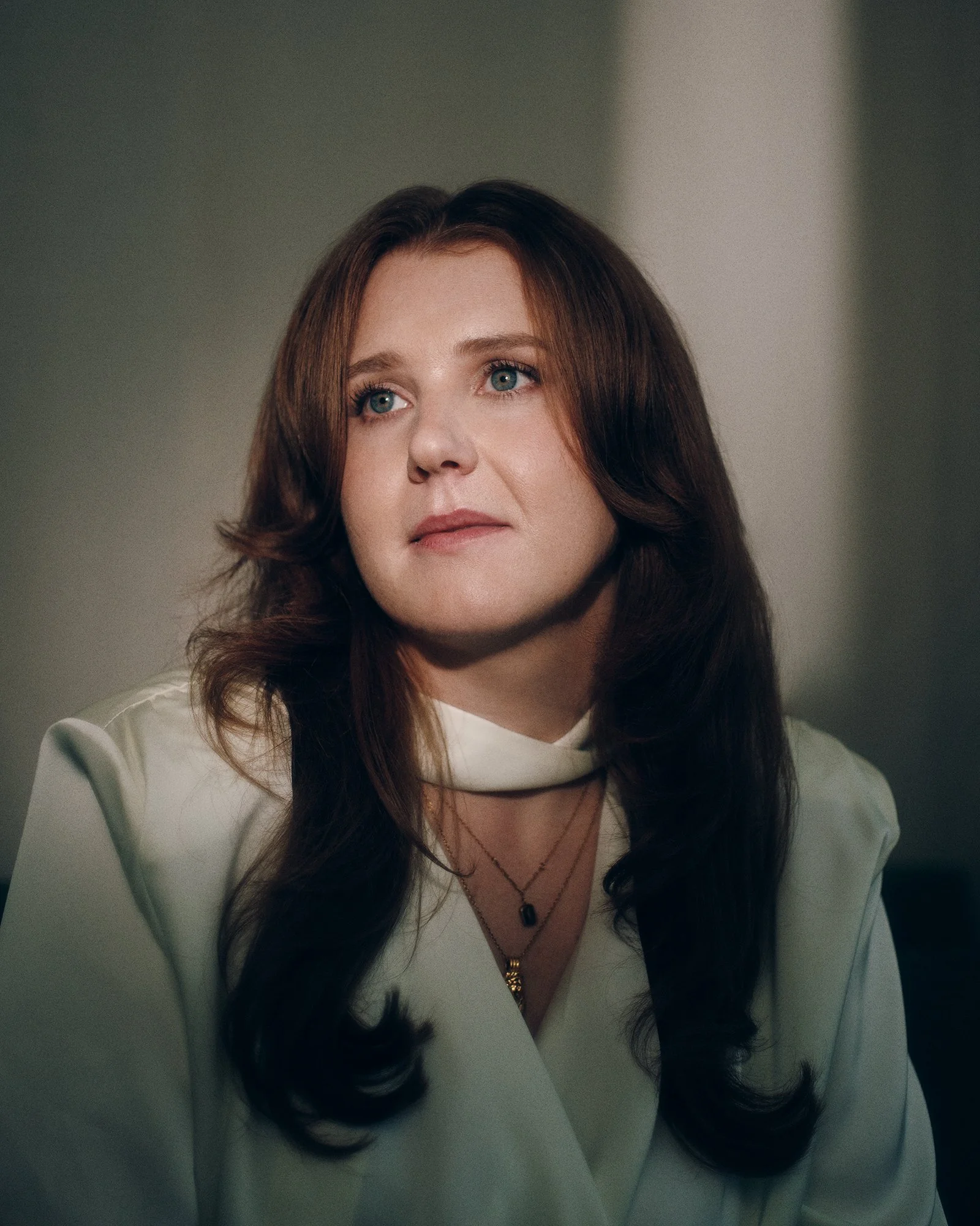 Portrait of a woman with long, dark hair and blue eyes, wearing a white top and layered necklaces, looking slightly upward, with a soft, neutral background.