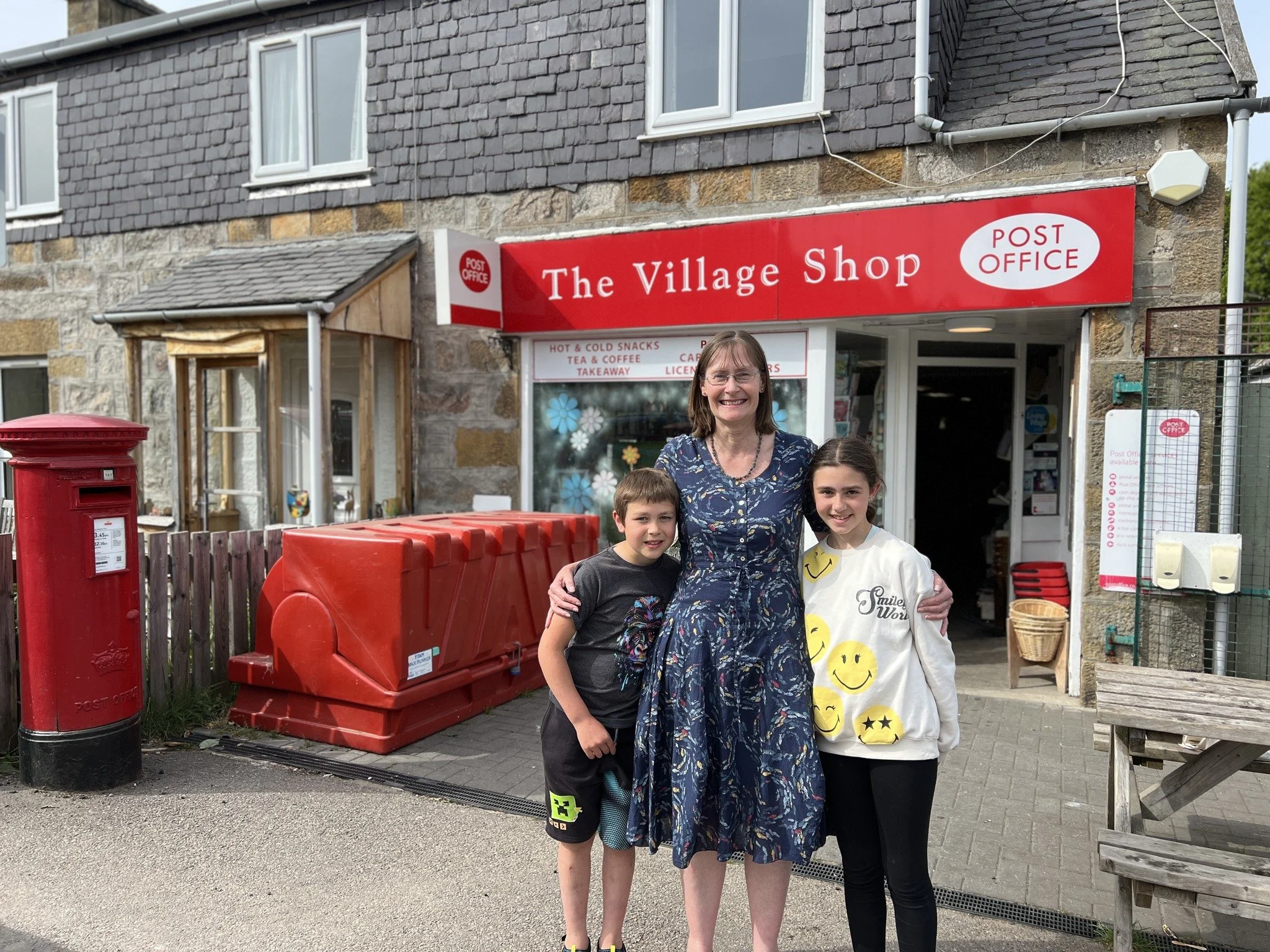 A woman and two children standing outside a shop called 'The Village Shop' with a red post office sign. The woman is in the middle, smiling, wearing a blue patterned dress, with her arms around a boy and a girl on either side. The boy is wearing a dark T-shirt and shorts, and the girl is wearing a white sweatshirt with smiley faces.