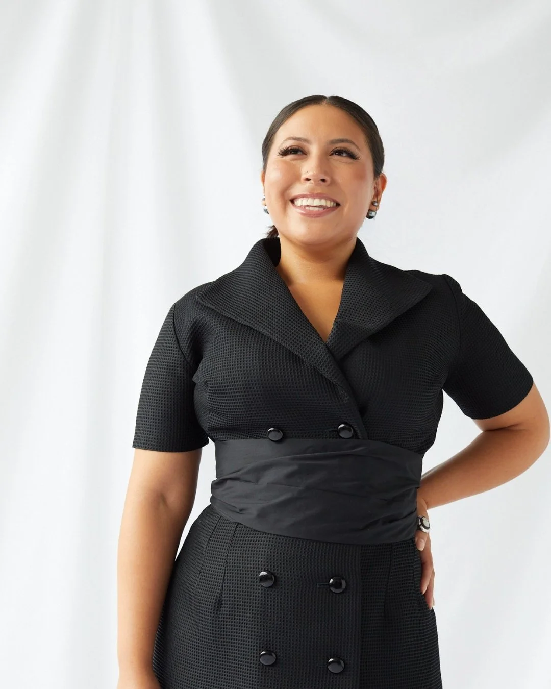 A woman wearing a black, textured dress with a wide collar, standing against a white background, smiling and looking to her right.