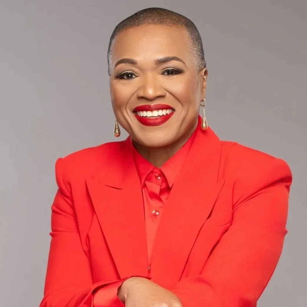A headshot of Dana Cox, a Black woman with a shaved head wearing bright red lipstick and a suit to match