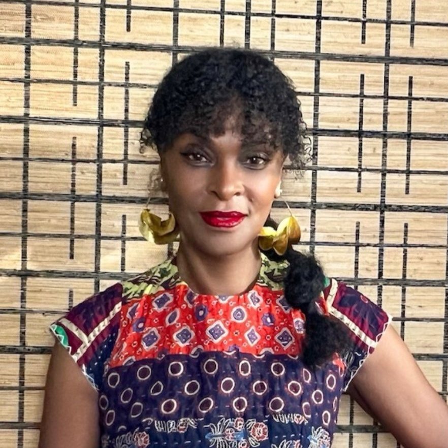 A headshot of Debra Da Costa, a woman with dark curly hair pulled back in a look ponytail wearing large gold earrings and a colorful, multi-print top