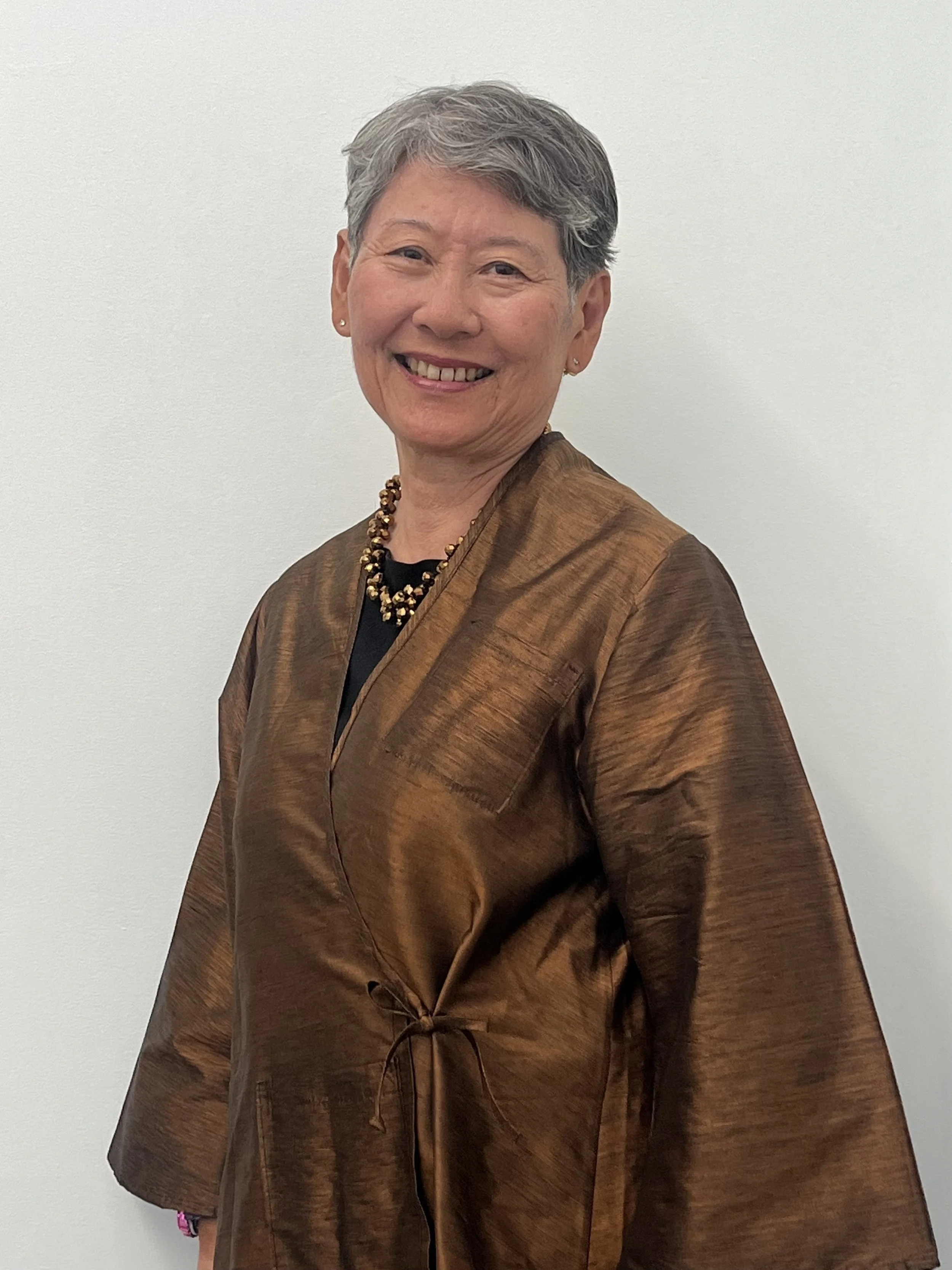 Pat Kozu, an older Japanese woman with short gray hair smiles in front of a blank wall while wearing a brown wrap top