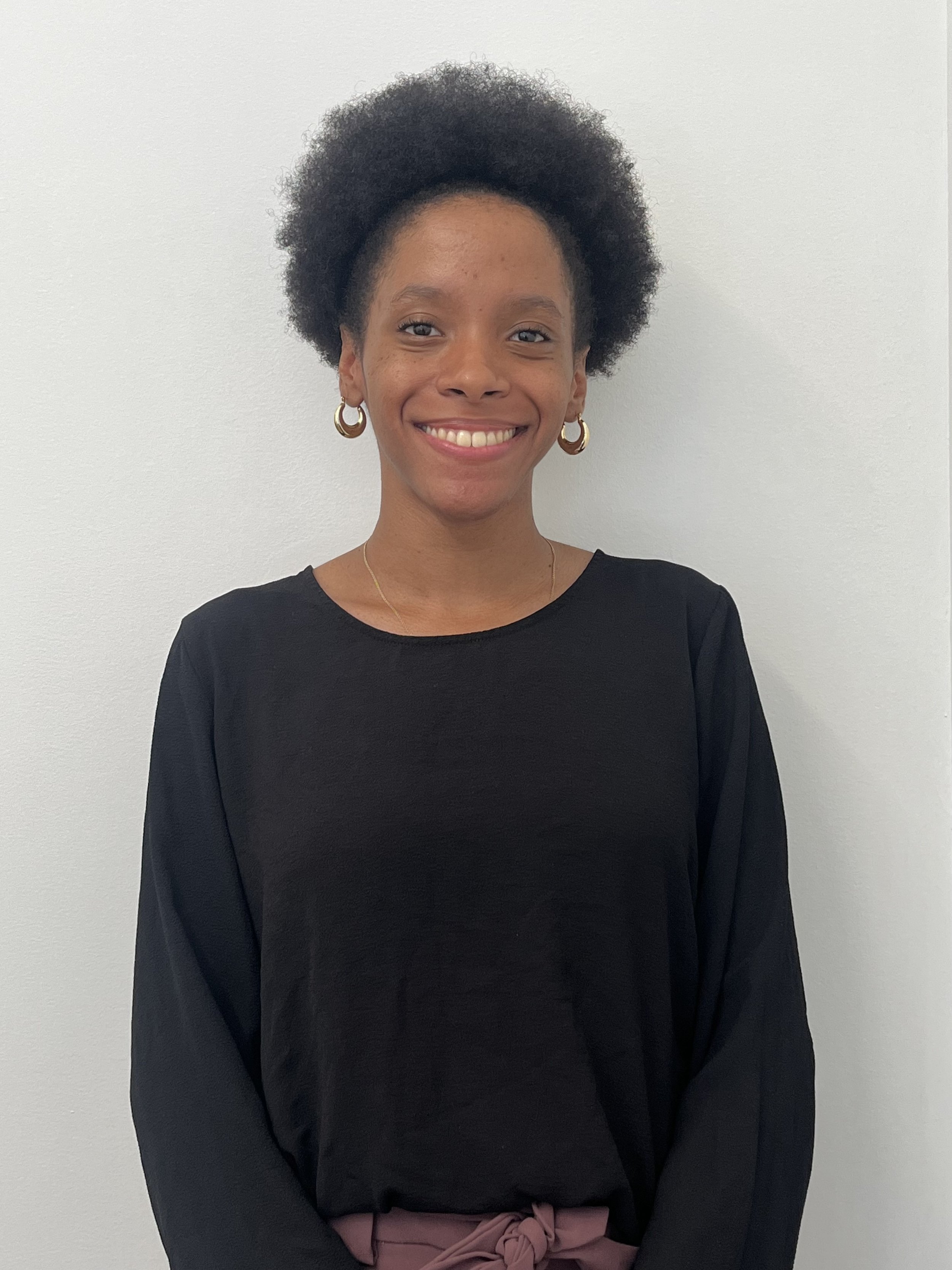 Doris Monsac, a young Black woman with natural hair smiles in front of a blank wall while wearing a black top