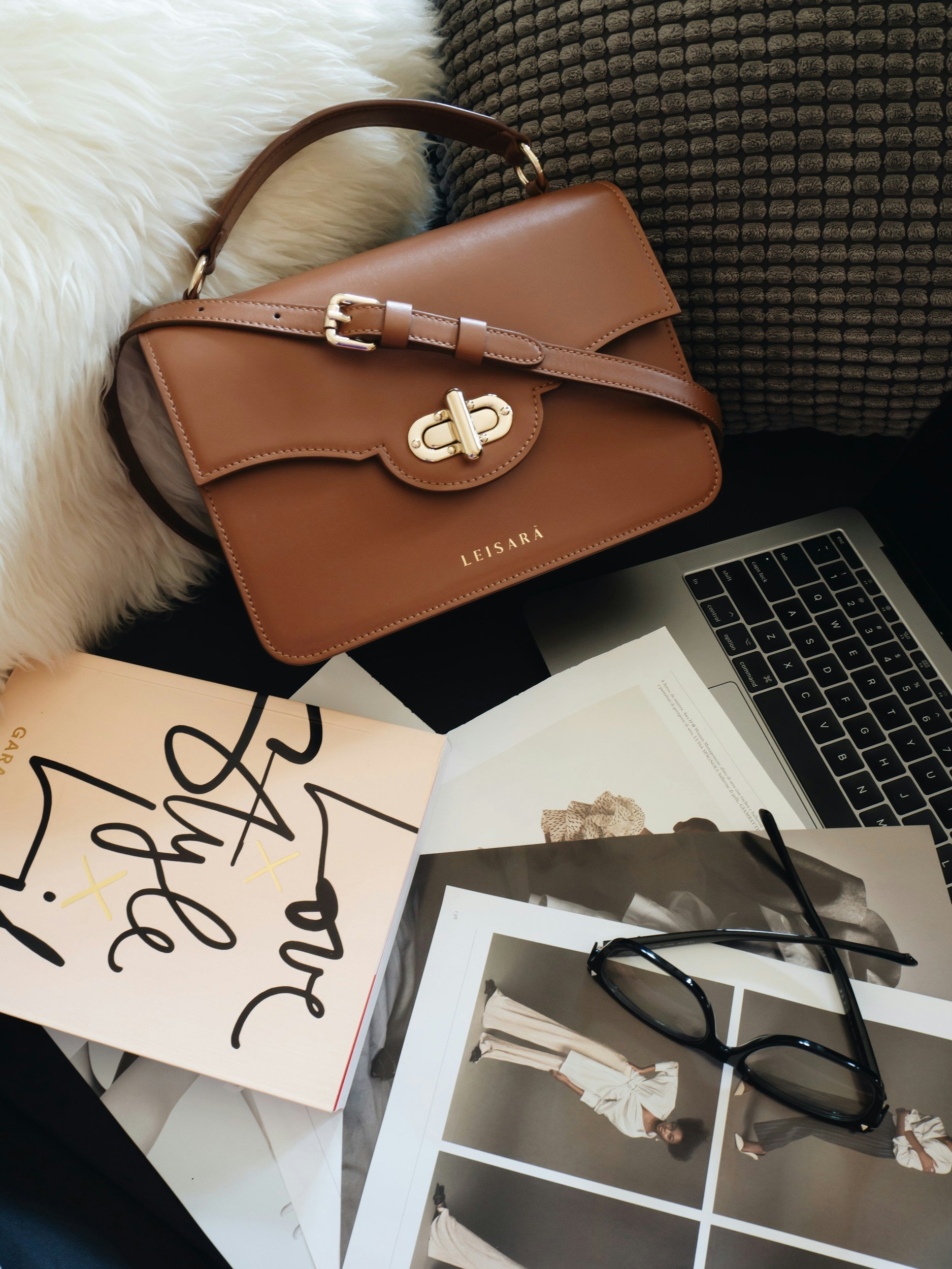 A brown leather handbag resting on a table, surrounded by fashion photographs, glasses, and printed materials.