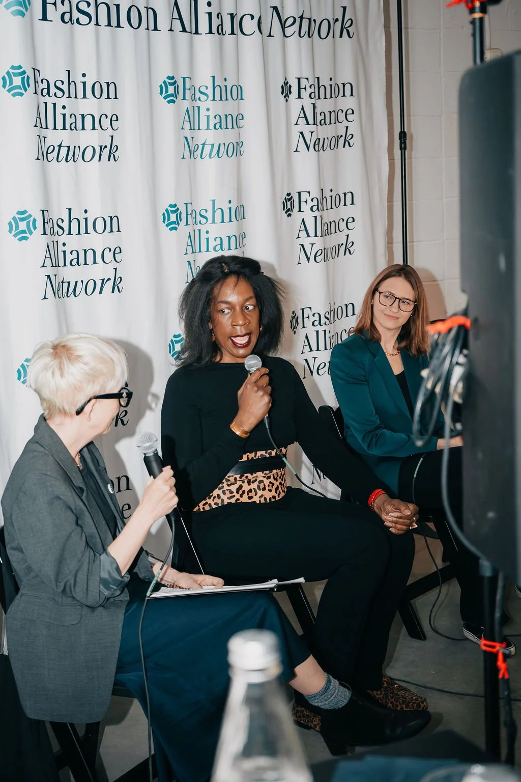 a woman holding a microphone with two other women on either side. All sitting in front of a backdrop that reads "Fashion Alliance Network"