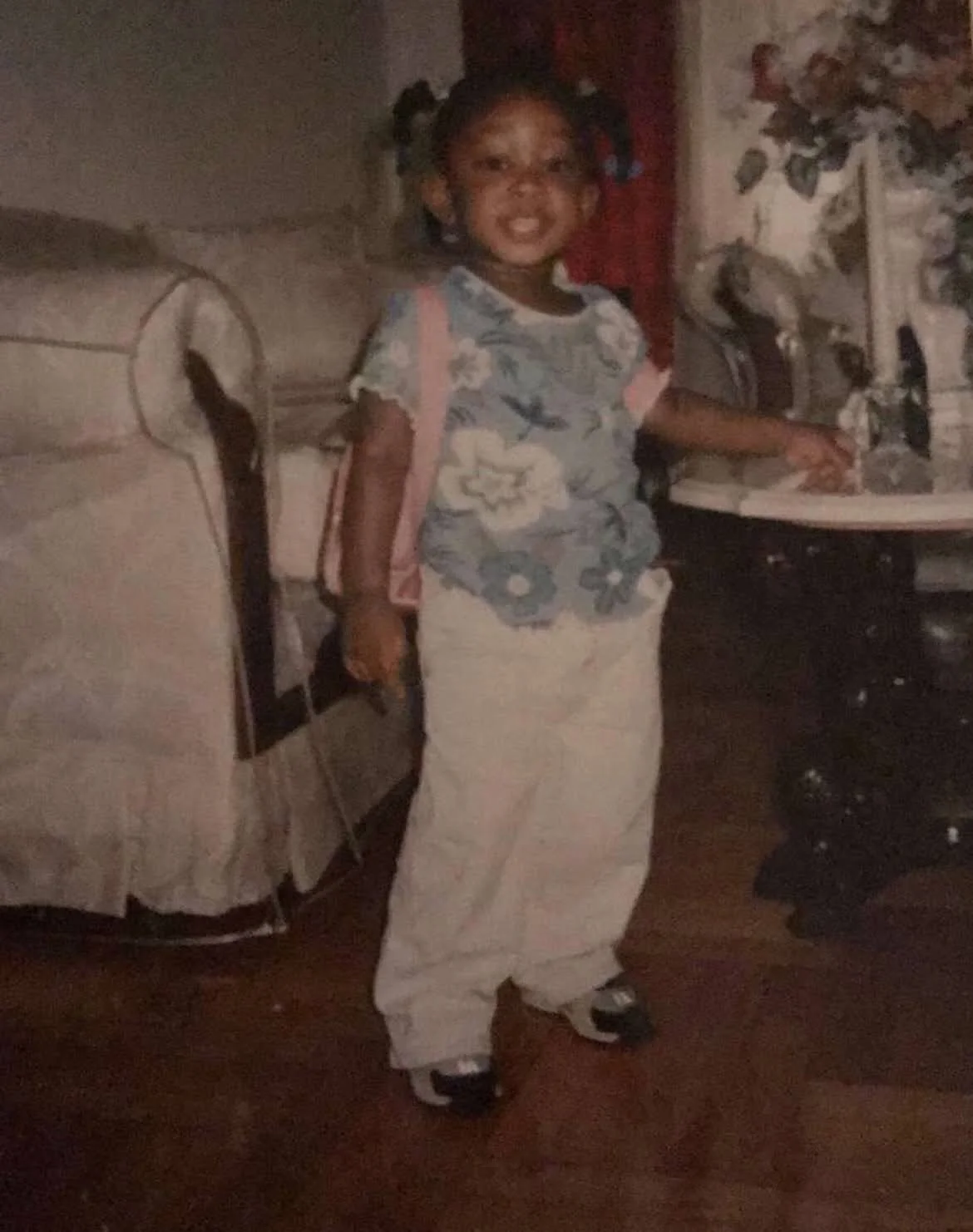 a young Black female child wearing a blue floral top with khaki pants, sneakers, and a pink backpack stands in a living room