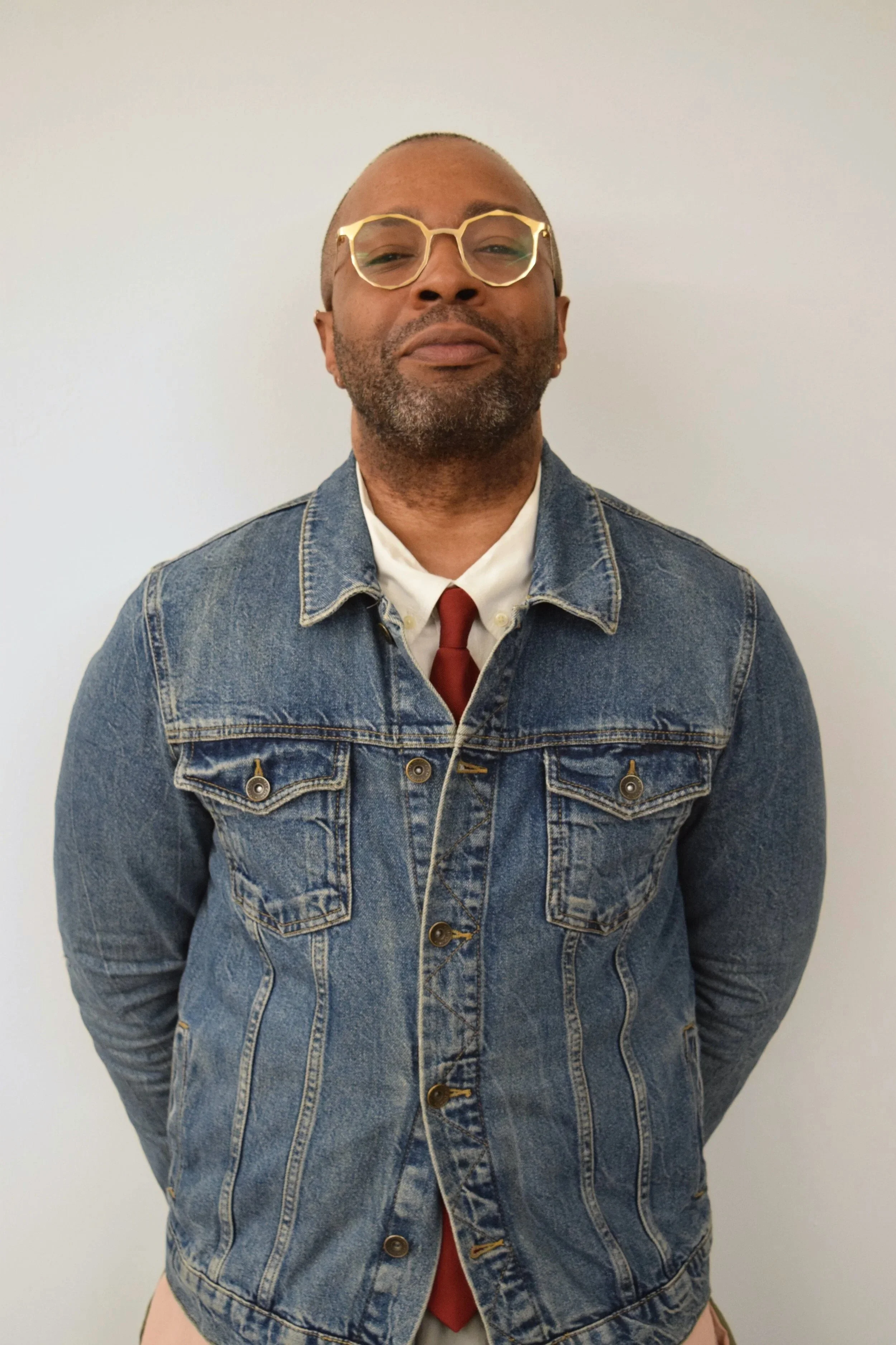 a Black man with very short hair wearing gold glasses, a collared shirt and tie under a denim jacket