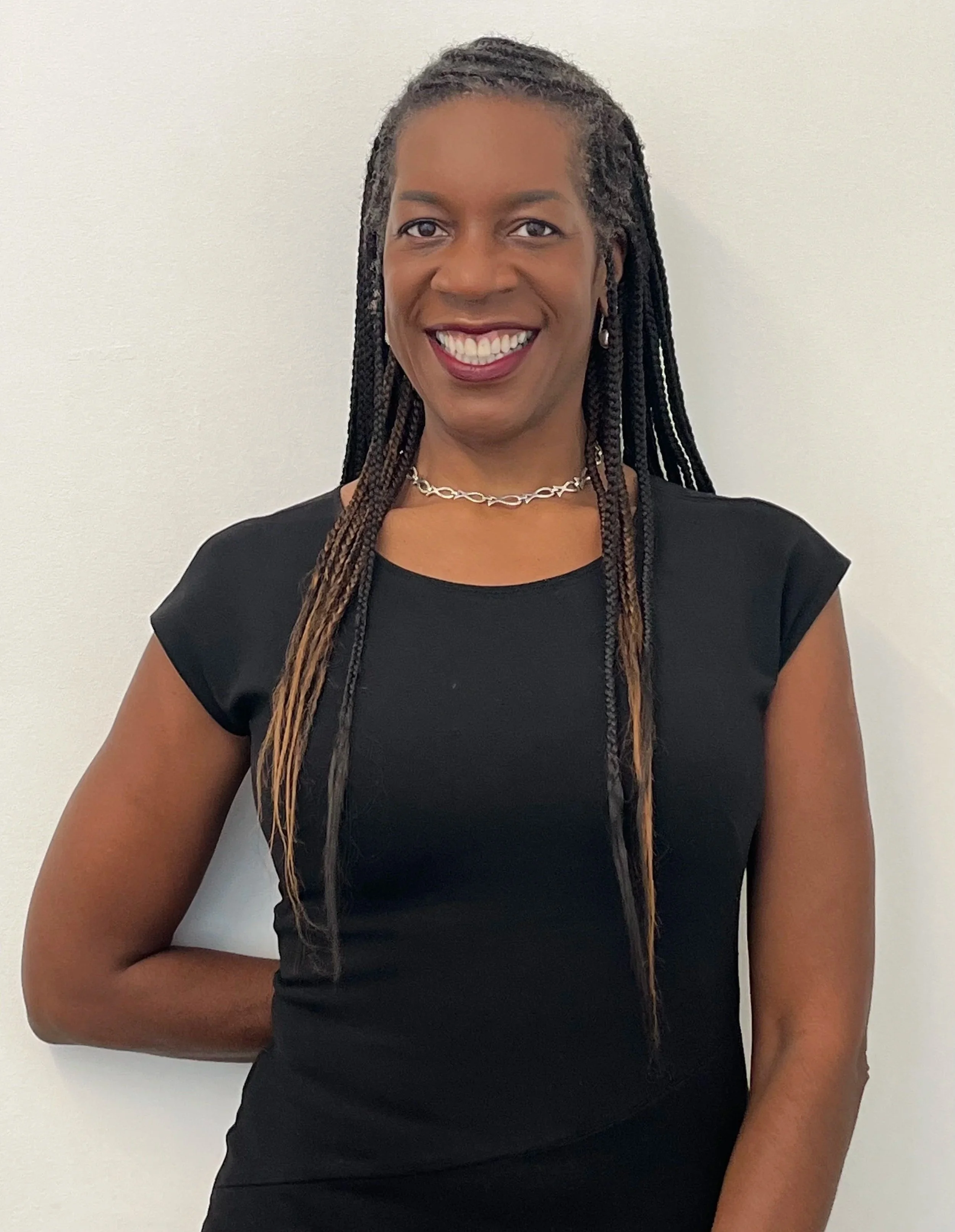 A woman with long braided hair, wearing a black short-sleeve top, silver necklace, and stud earrings, smiling and standing against a plain white wall.