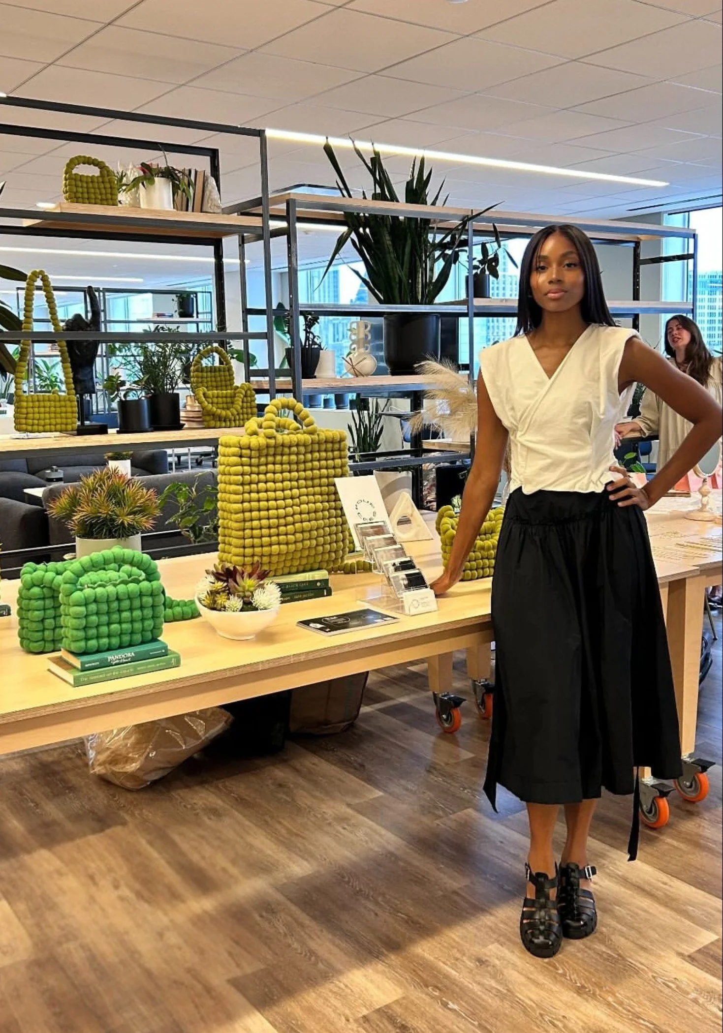 A woman with long dark hair, wearing a beige sleeveless top and black skirt, stands in a modern retail store surrounded by decorative handbags and plants on display shelves. She has her hand on her hip and is wearing chunky black sandals. Another woman can be seen in the background smiling.