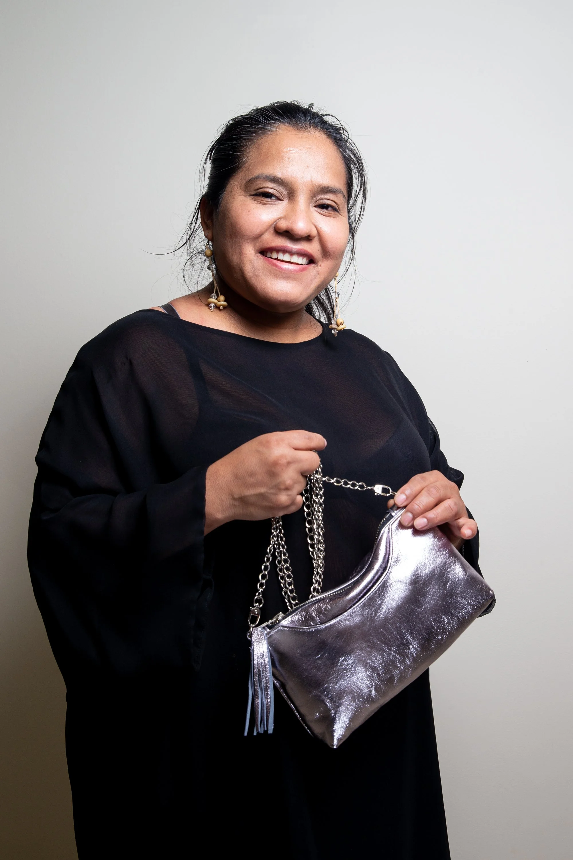 A woman with dark hair, wearing earrings, holding a shiny silver clutch purse, standing against a plain light gray wall, smiling at the camera.