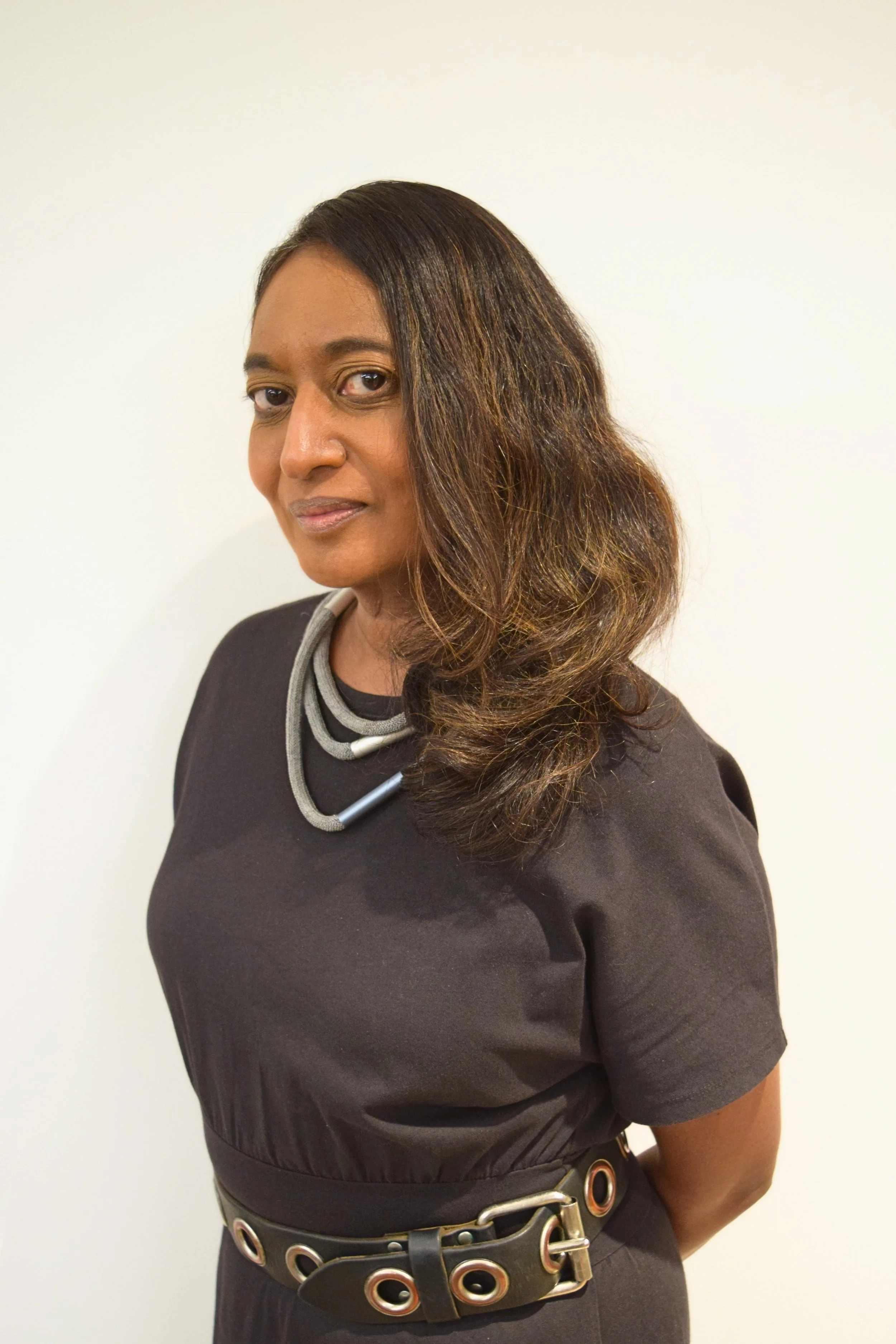 a woman with chest-length brown hair wearing a black dress with a layered silver necklace and rivet belt