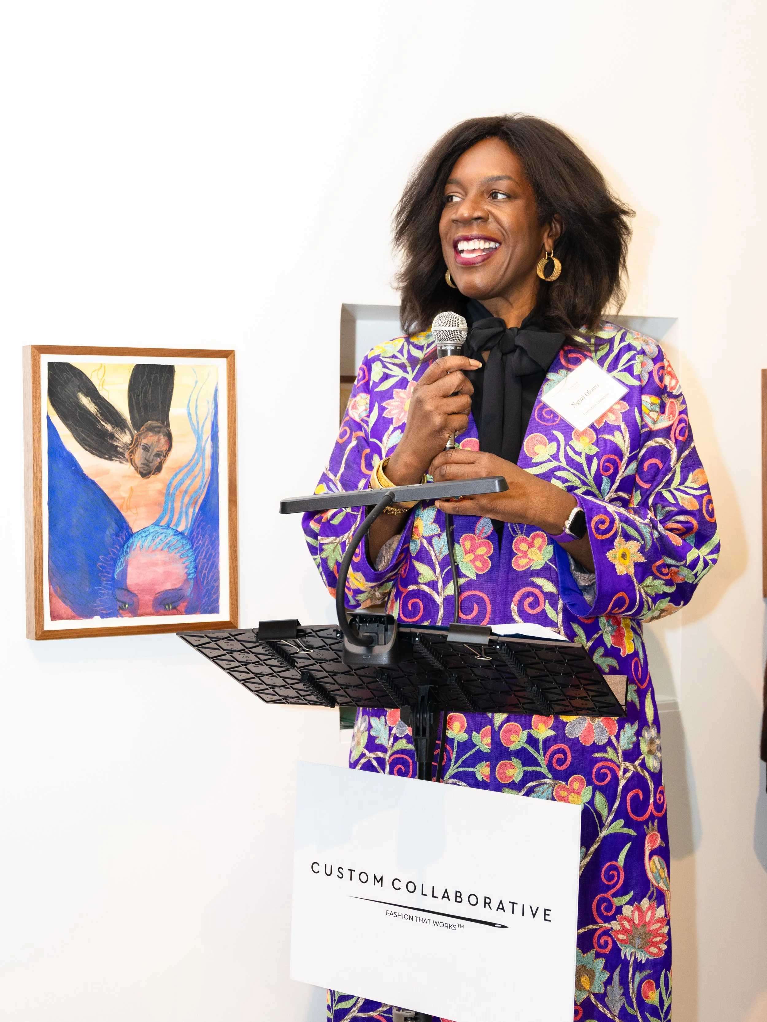 Ngozi Okaro wearing a printed purple jacket stands at a podium with a mounted Custom Collaborative sign while speaking into a microphone 
