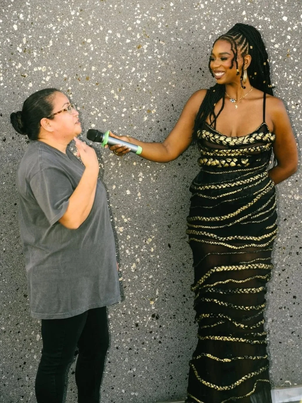 Jazzmine, a tall Black woman with braided hair wearing a strapless black and gold dress, holds out a microphone for Rosa, a Latina woman with hair pulled into a bun wearing glasses, a gray T-shirt, and black pants