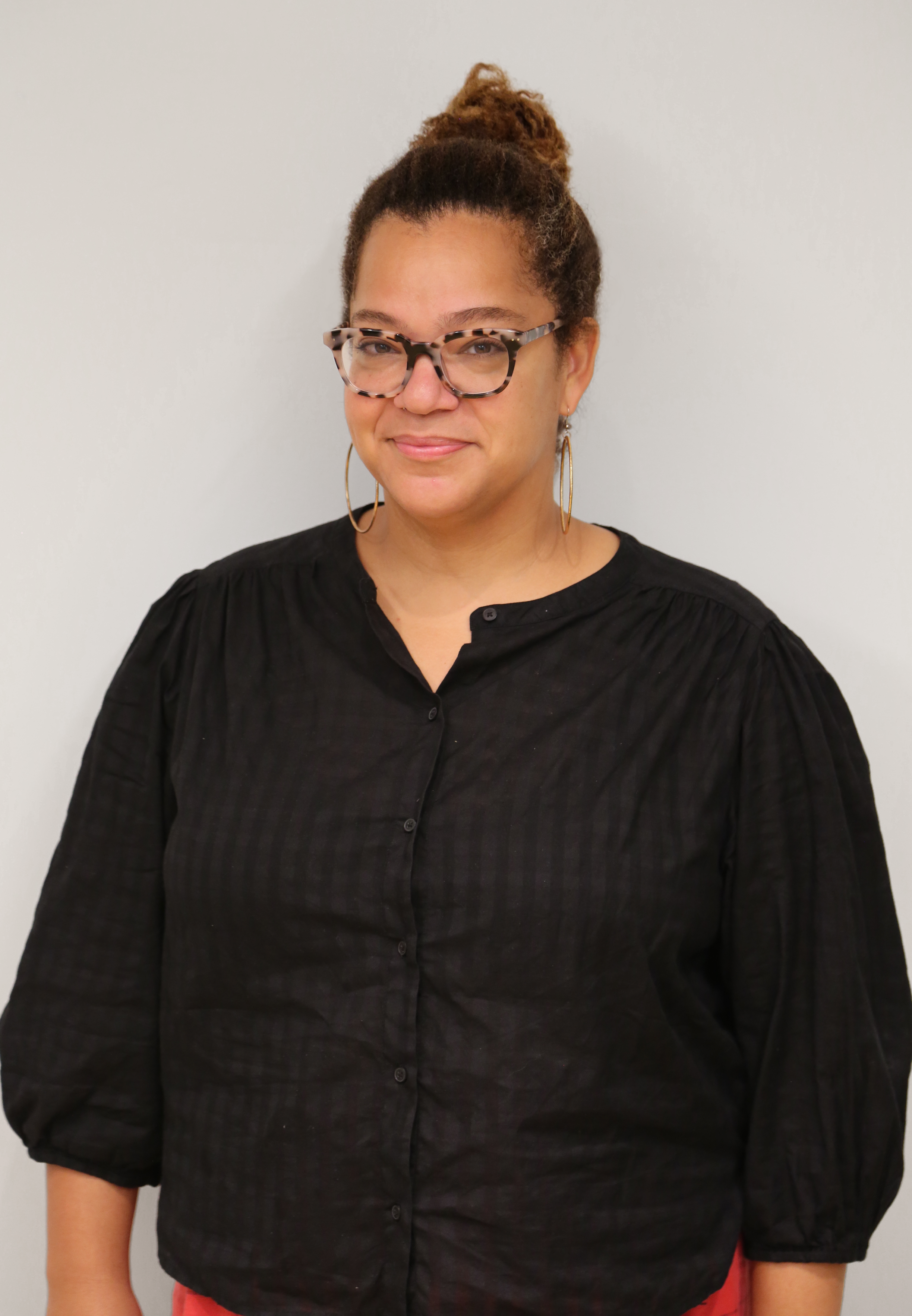 A woman wearing glasses with a patterned frame, hoop earrings, and a black button-up shirt with ruched sleeves, standing against a plain light background.