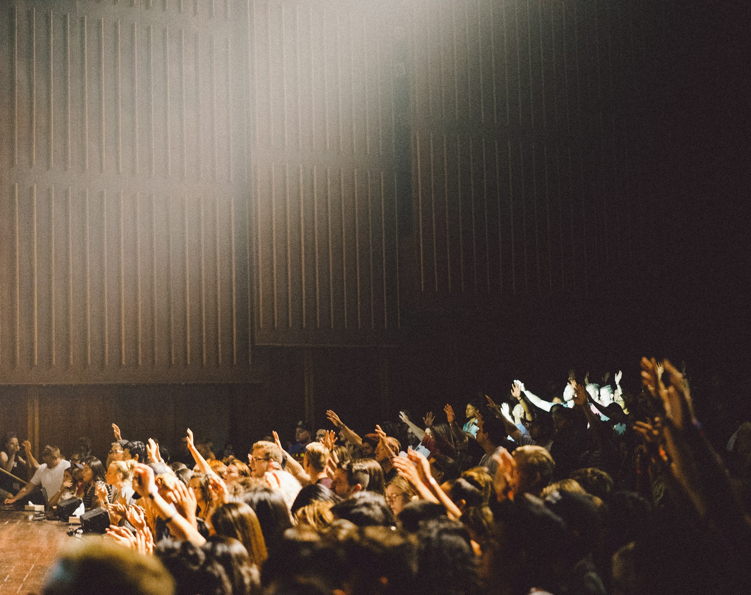 Crowd of people in an auditorium with many raising their hands, in a dimly lit room with wooden panel walls.