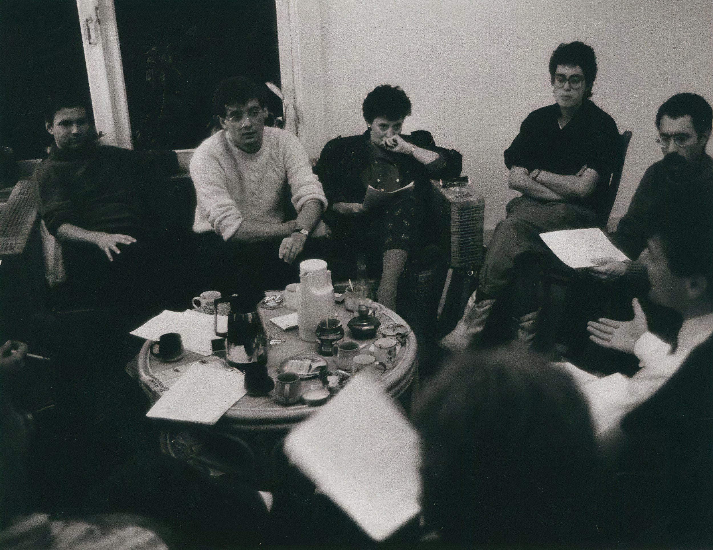 Black and white photo of six people sitting around a coffee table in a room, some with notebooks and drinks, engaging in a discussion or meeting.