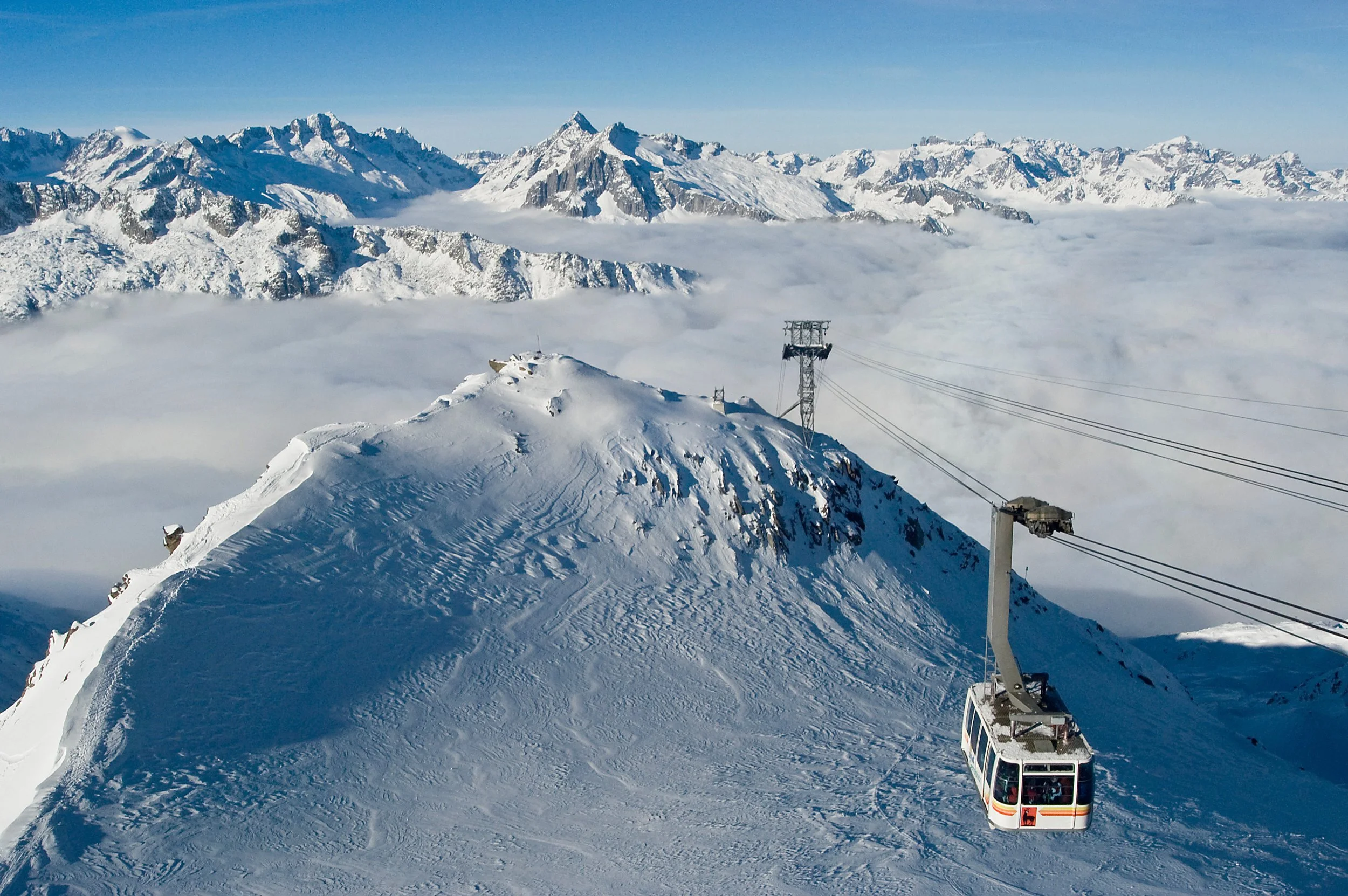Snow-covered mountain peak with a cable car ascending, surrounded by snow-capped mountains and clouds in the distance.